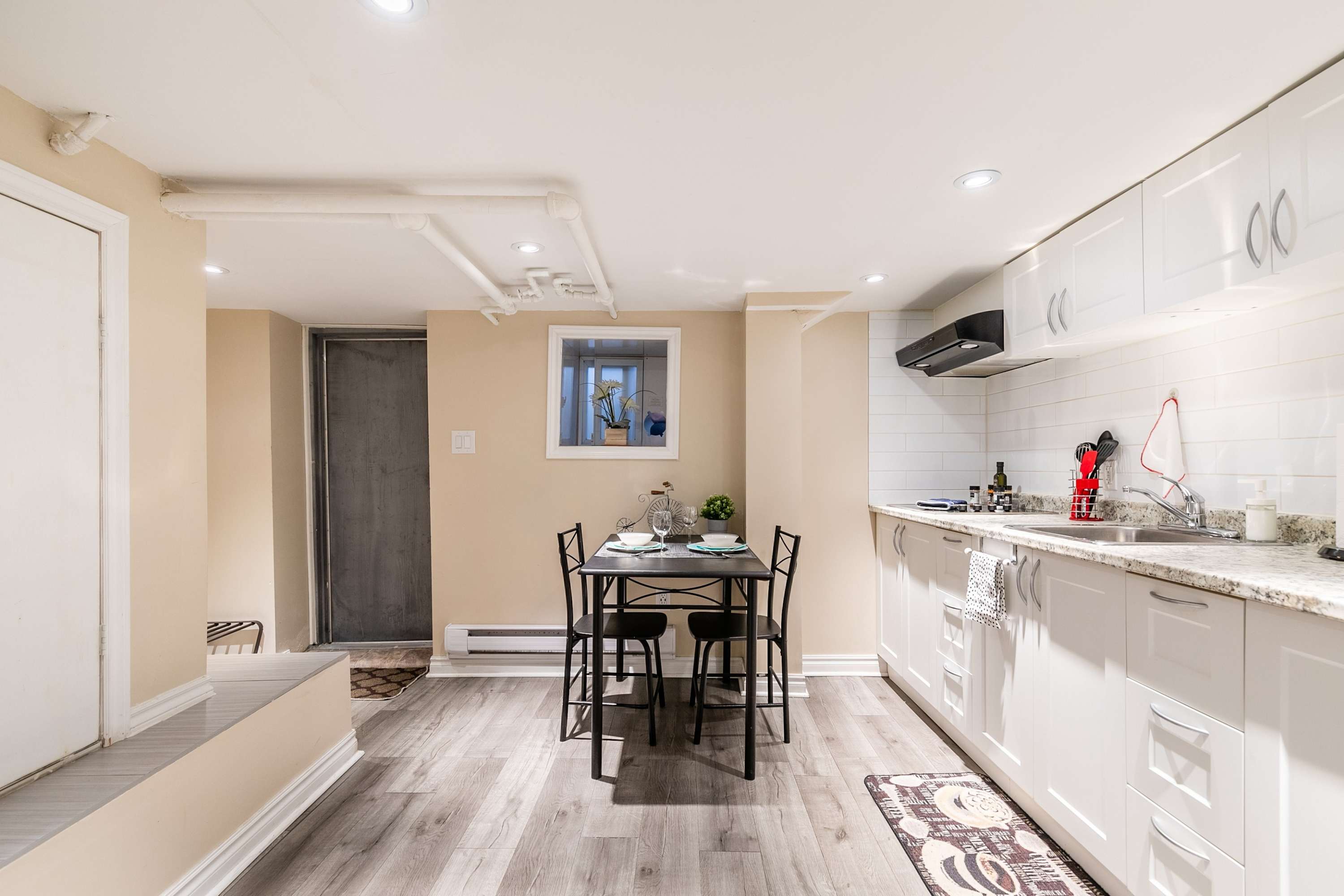 A wide shot of the kitchen and dining area, showcasing white shaker cabinets, light granite countertops, and a black dining table with chairs. The space features light wood-look flooring and recessed lighting, with exposed pipes on the ceiling.