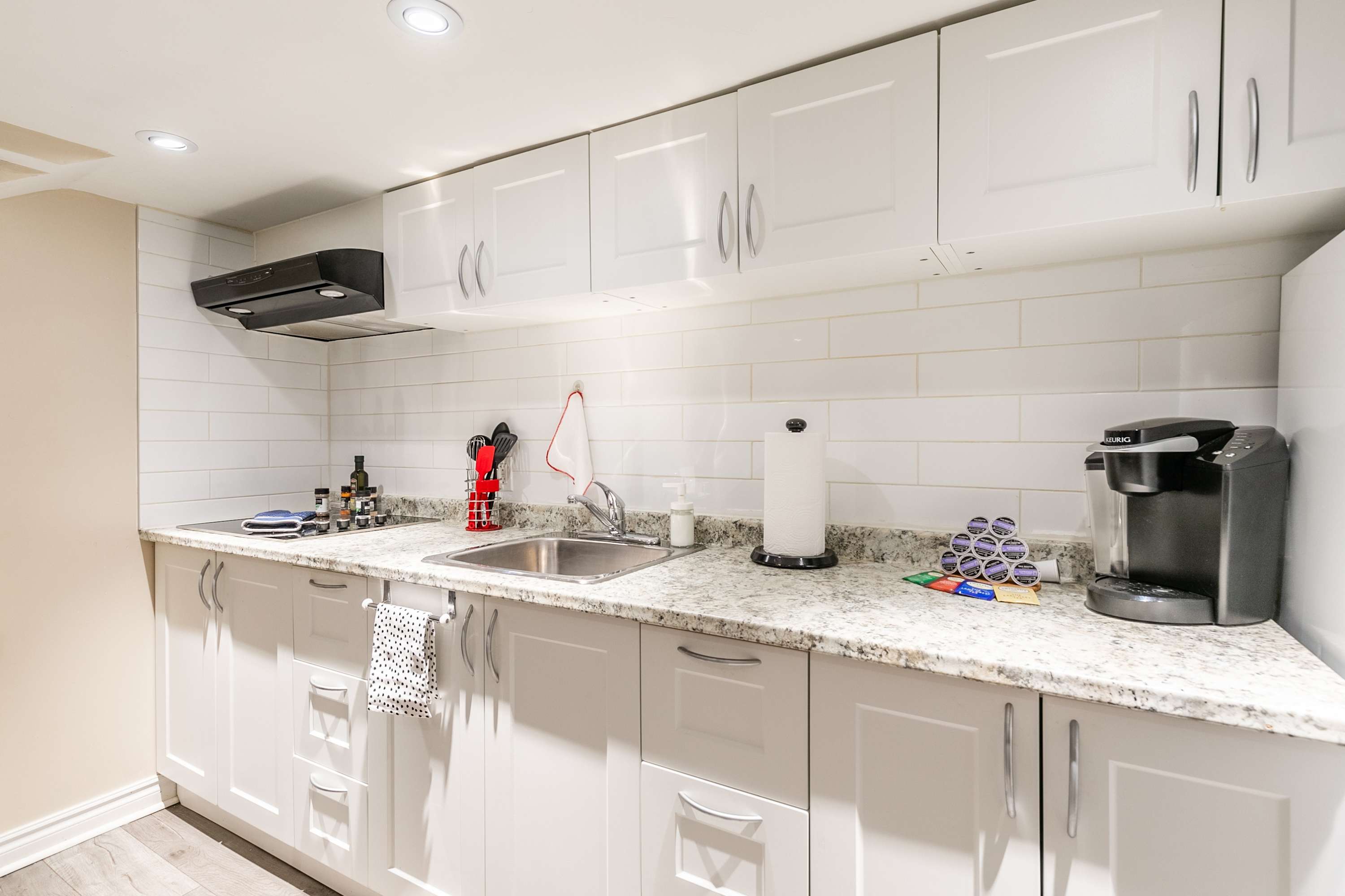 A detailed view of the kitchen countertop and cabinets, highlighting the white shaker style, light granite surface, and white subway tile backsplash. A stainless steel sink, Keurig coffee maker, and various kitchen utensils are neatly arranged.