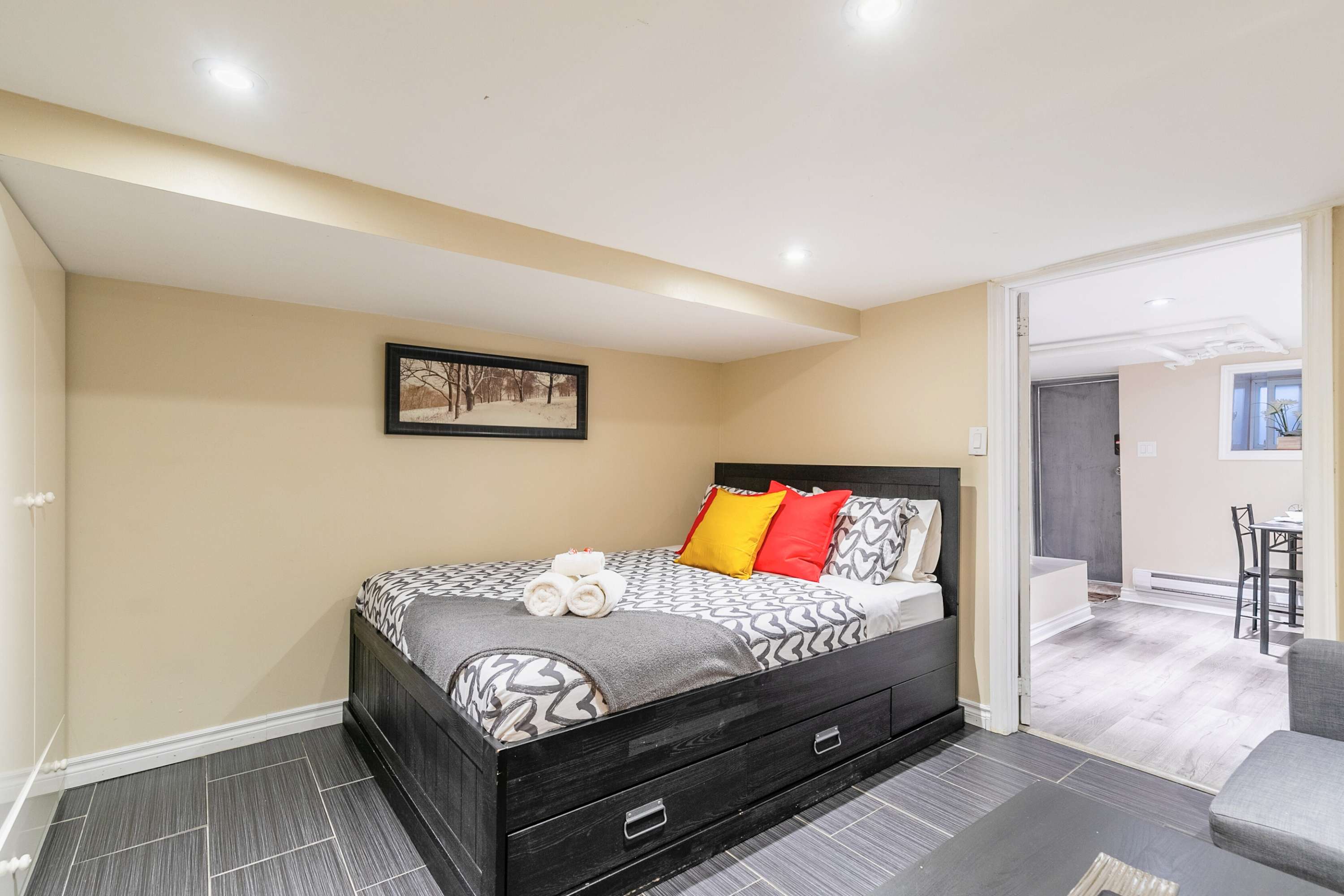 Inviting bedroom with a black bed frame, a patterned grey and white duvet, and vibrant red and yellow throw pillows, accented by neatly rolled white towels. A framed winter landscape hangs above the bed on the light beige wall, with dark grey tiled flooring throughout.