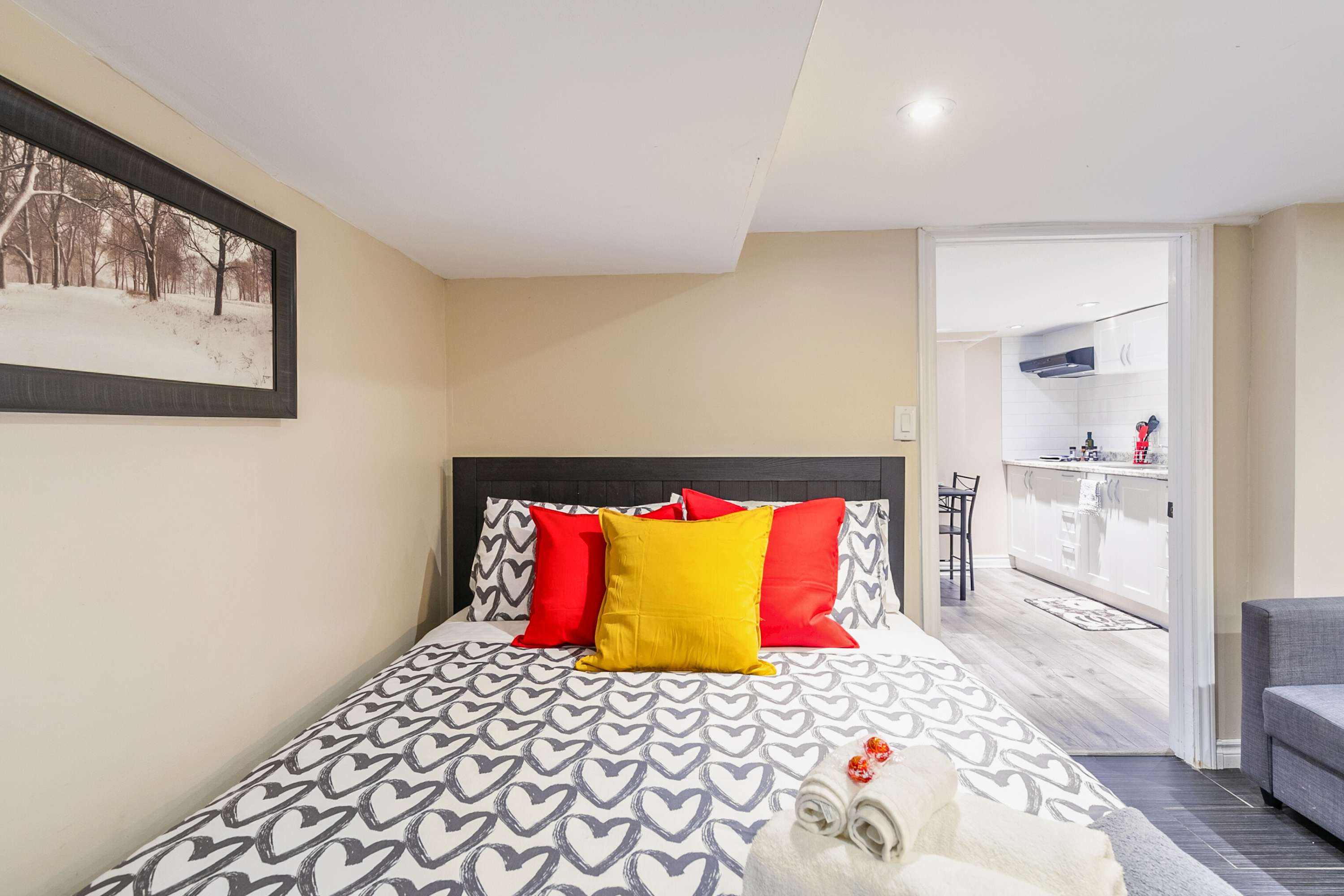 Close-up view of the bed in the bedroom, featuring a black headboard, patterned duvet, and bright red and yellow pillows, with neatly folded white towels at the foot. A doorway on the right opens to reveal a glimpse of the kitchen area beyond.