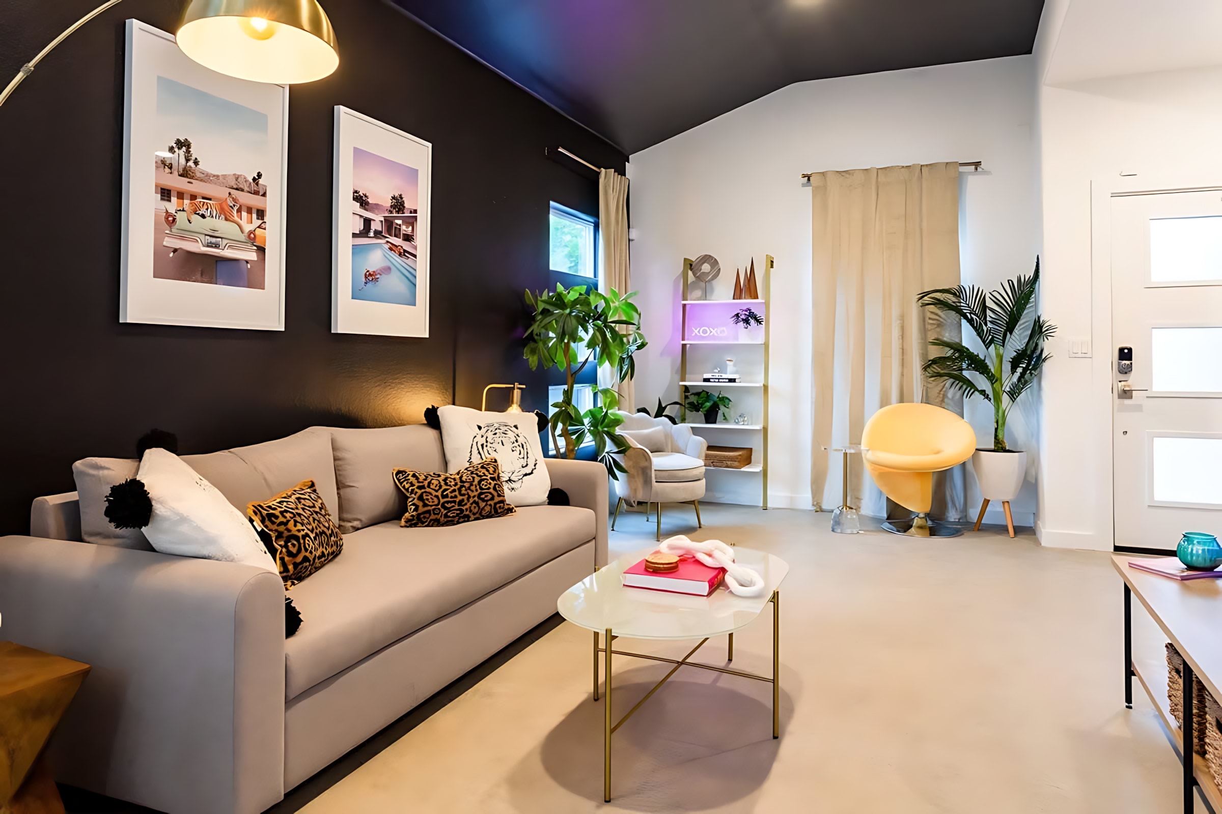 A stylish living room with a light gray sofa, a glass coffee table, and a gold arc lamp, complemented by a white shelving unit displaying decor and a neon "XOXO" sign. Large windows with light-colored drapes and a variety of indoor plants add to the bright, inviting atmosphere.