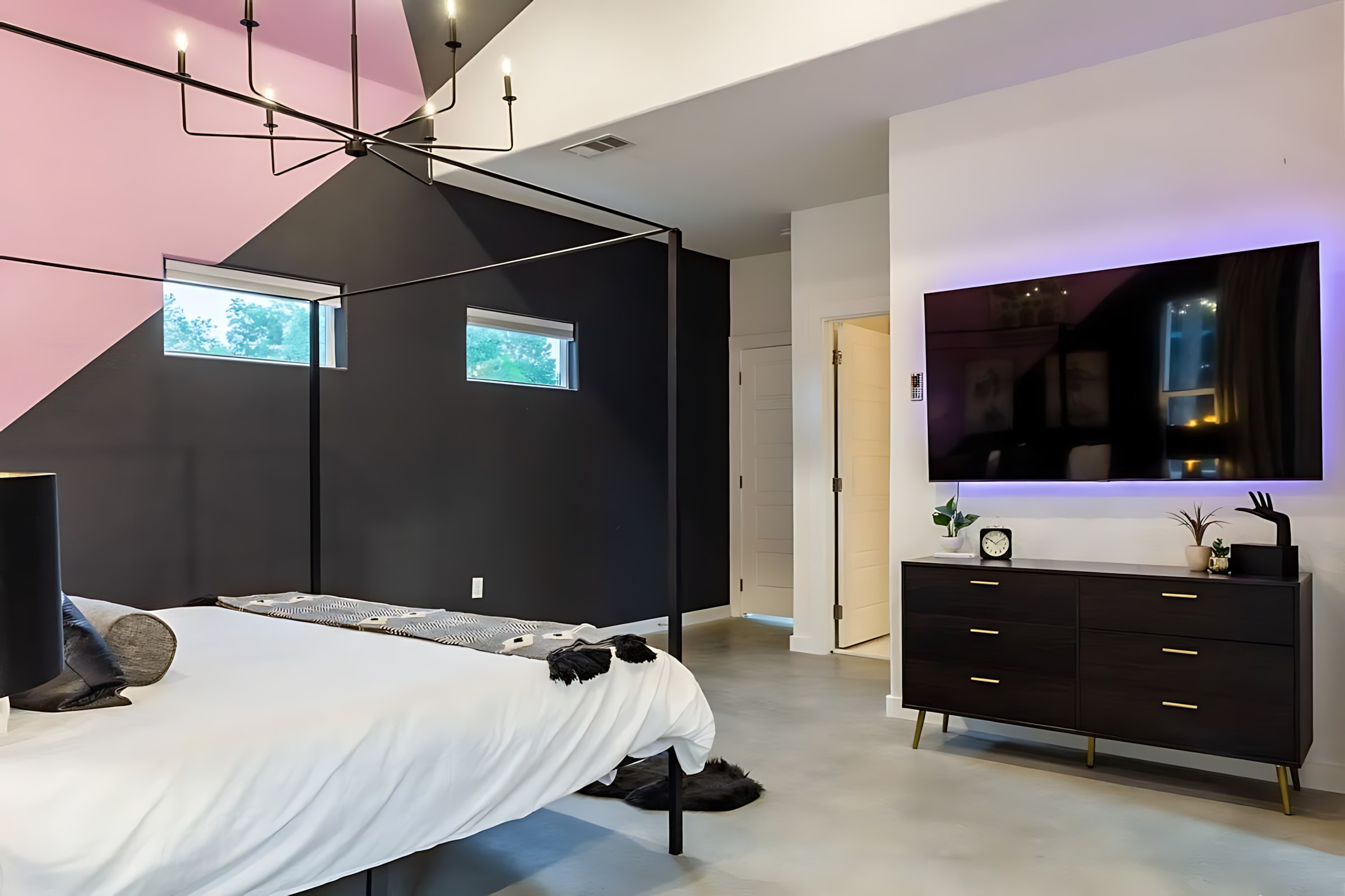 A wide shot of the modern bedroom with a large bed, black and pink geometric walls, and a wall-mounted TV above a dark wood dresser with gold accents, illuminated by a contemporary chandelier.