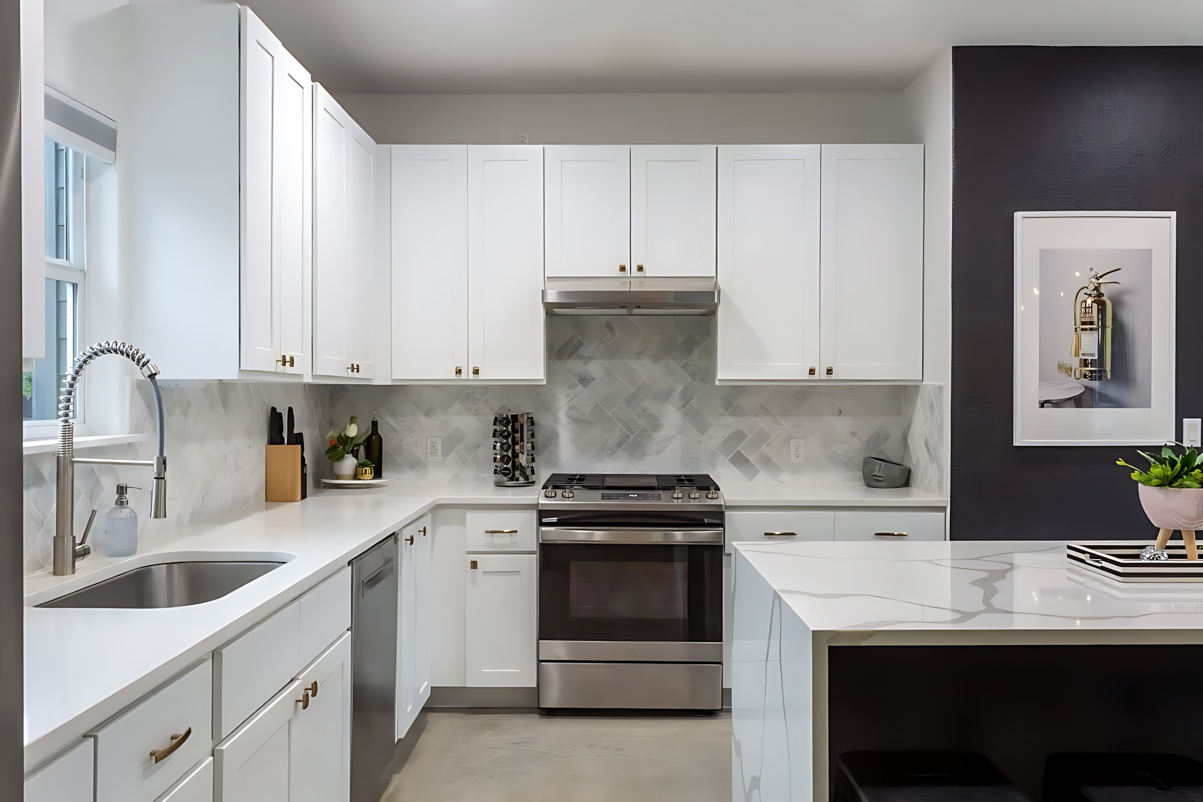 A modern kitchen with white shaker cabinets, white quartz countertops, and a gray herringbone tile backsplash, featuring stainless steel appliances including a range and dishwasher. A large window above the sink provides natural light.