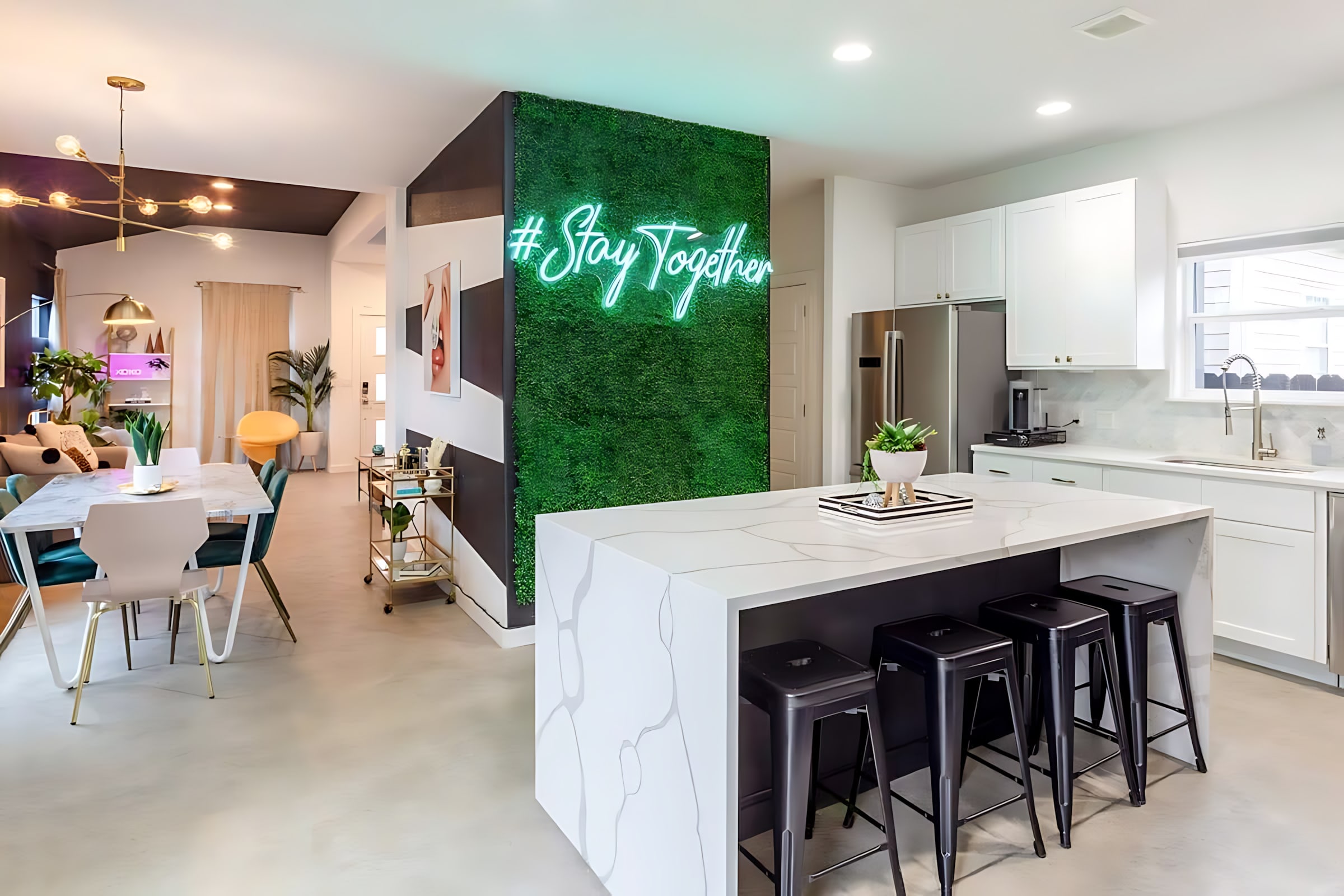 A modern kitchen boasts white cabinetry, stainless steel appliances, and a large island with a white quartz countertop and black bar stools. A vibrant green moss wall with a neon "#Stay Together" sign adds a unique focal point, leading into a dining area.