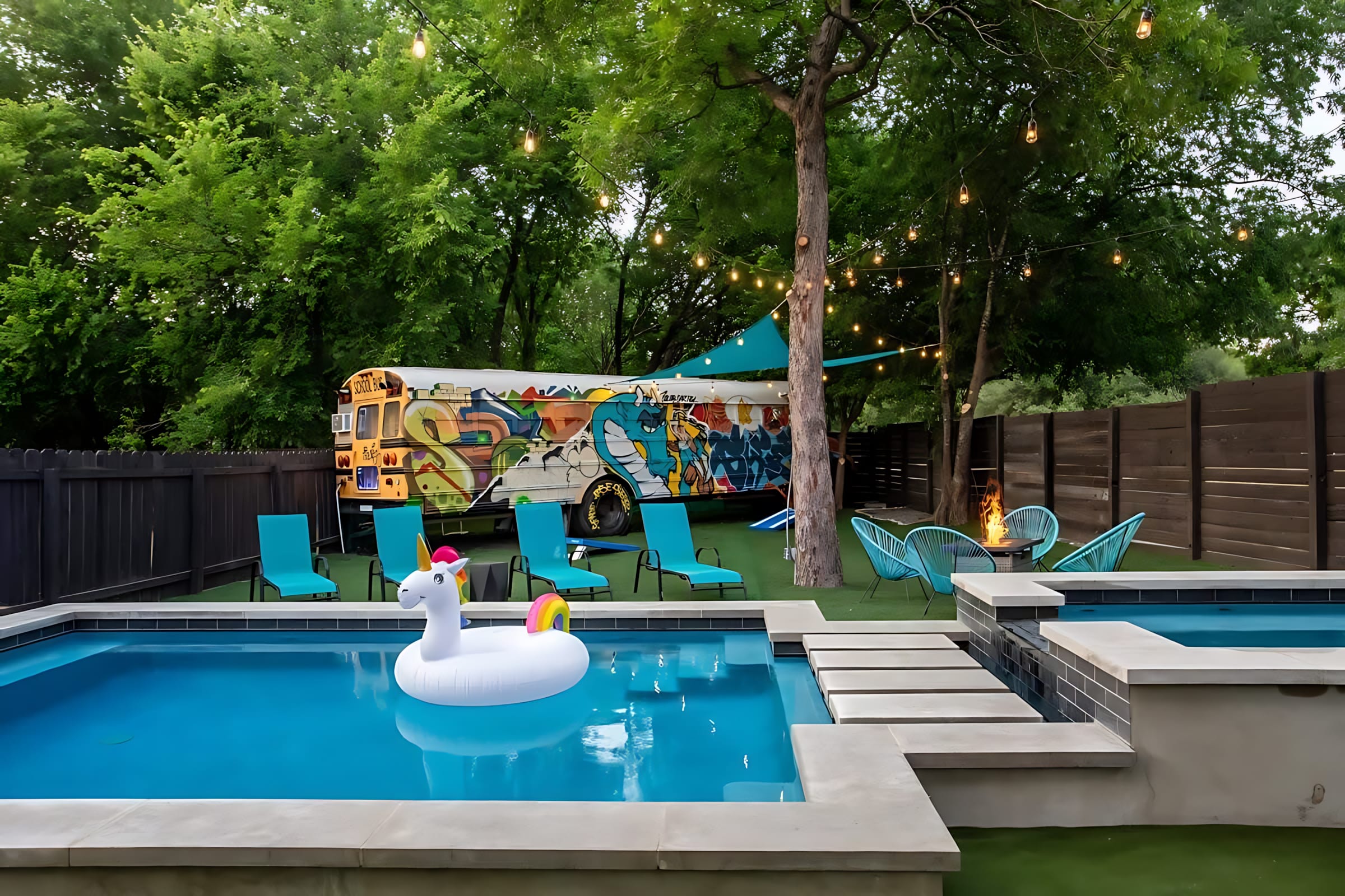 A vibrant outdoor space showcases a rectangular pool with a unicorn float and a hot tub, set on artificial turf. A large, graffiti-covered school bus is a prominent feature in the background, surrounded by mature trees and overhead string lights.
