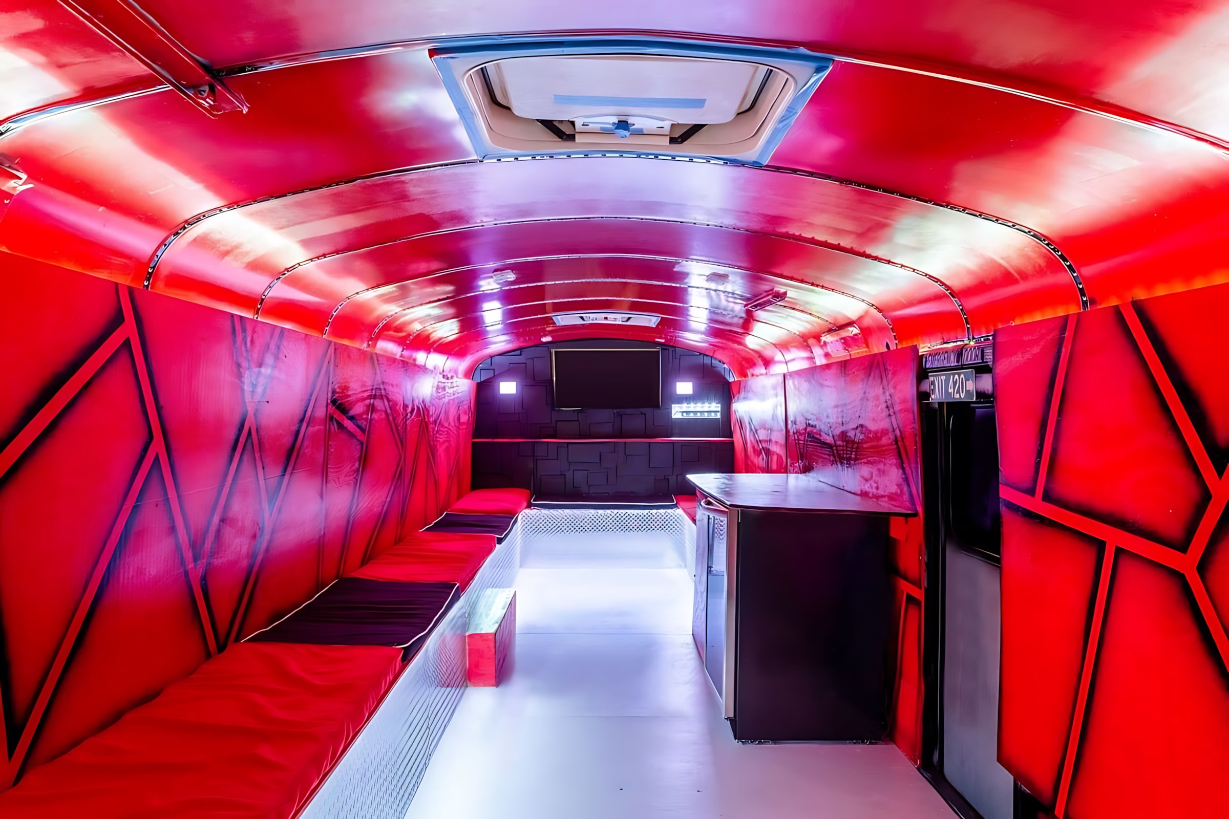 A long, narrow room with vibrant red LED lighting illuminating textured walls and ceiling, featuring black and red cushioned benches along both sides and a small bar area at the far end. The floor is white and reflective, enhancing the futuristic, club-like atmosphere.