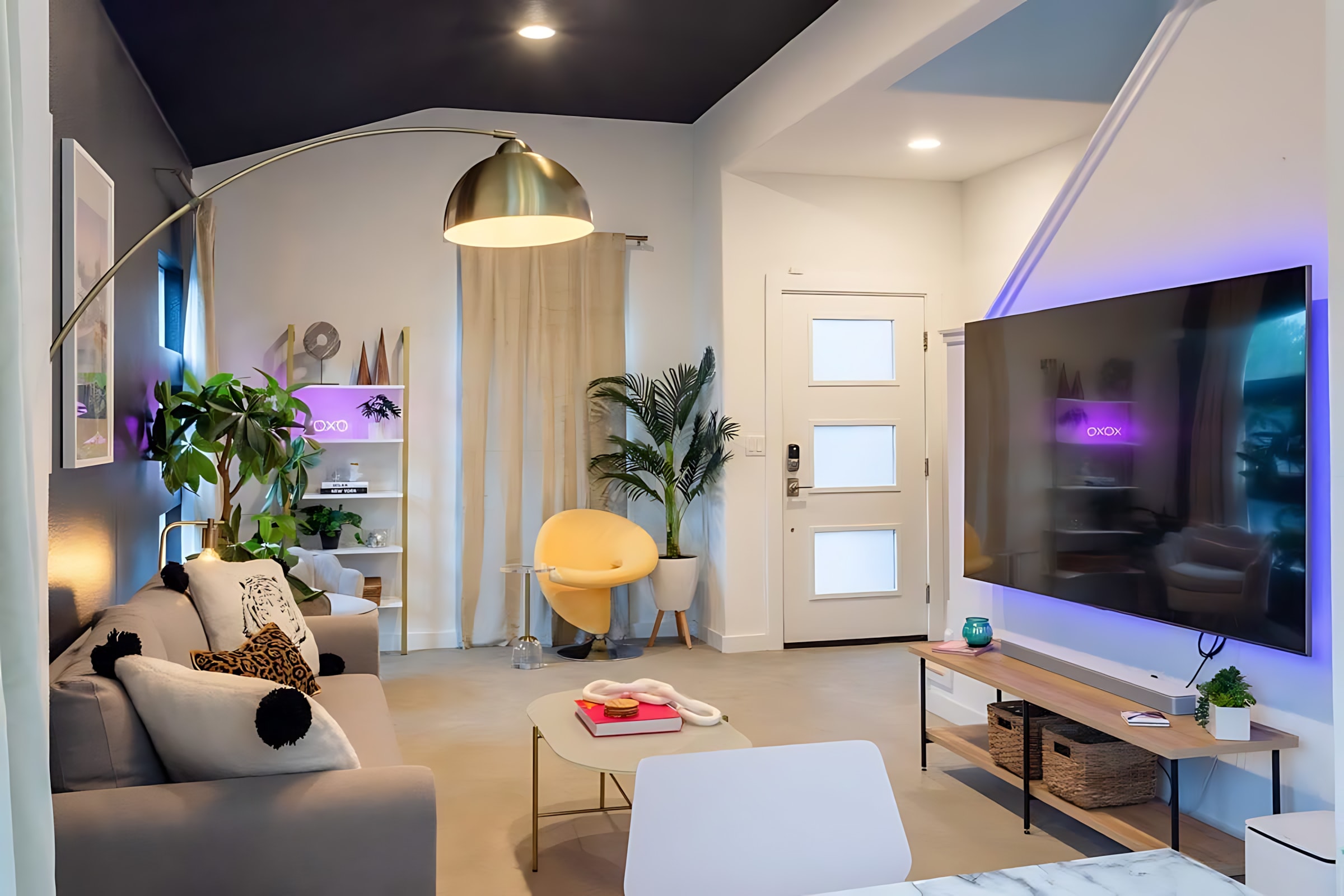 A contemporary living room features a grey sofa with decorative pillows, a large wall-mounted TV with purple backlighting, and a gold arc floor lamp. A yellow accent chair and various plants add pops of color and life to the space.