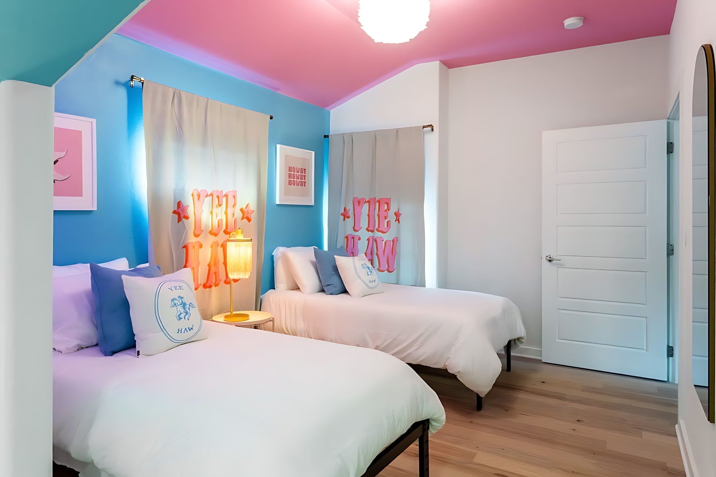 A colorful bedroom with three twin beds dressed in white, featuring blue accent pillows and a pink sloped ceiling. The room has bright blue walls with "YEE HAW" curtains and a white door.