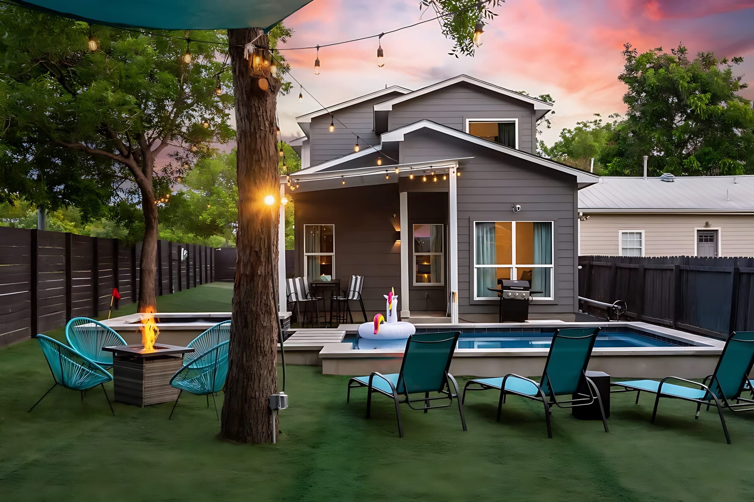 The back of a modern grey house overlooks a vibrant backyard with a pool, hot tub, and artificial turf. String lights illuminate the space, which includes lounge chairs, a fire pit, and a covered patio with outdoor dining furniture.