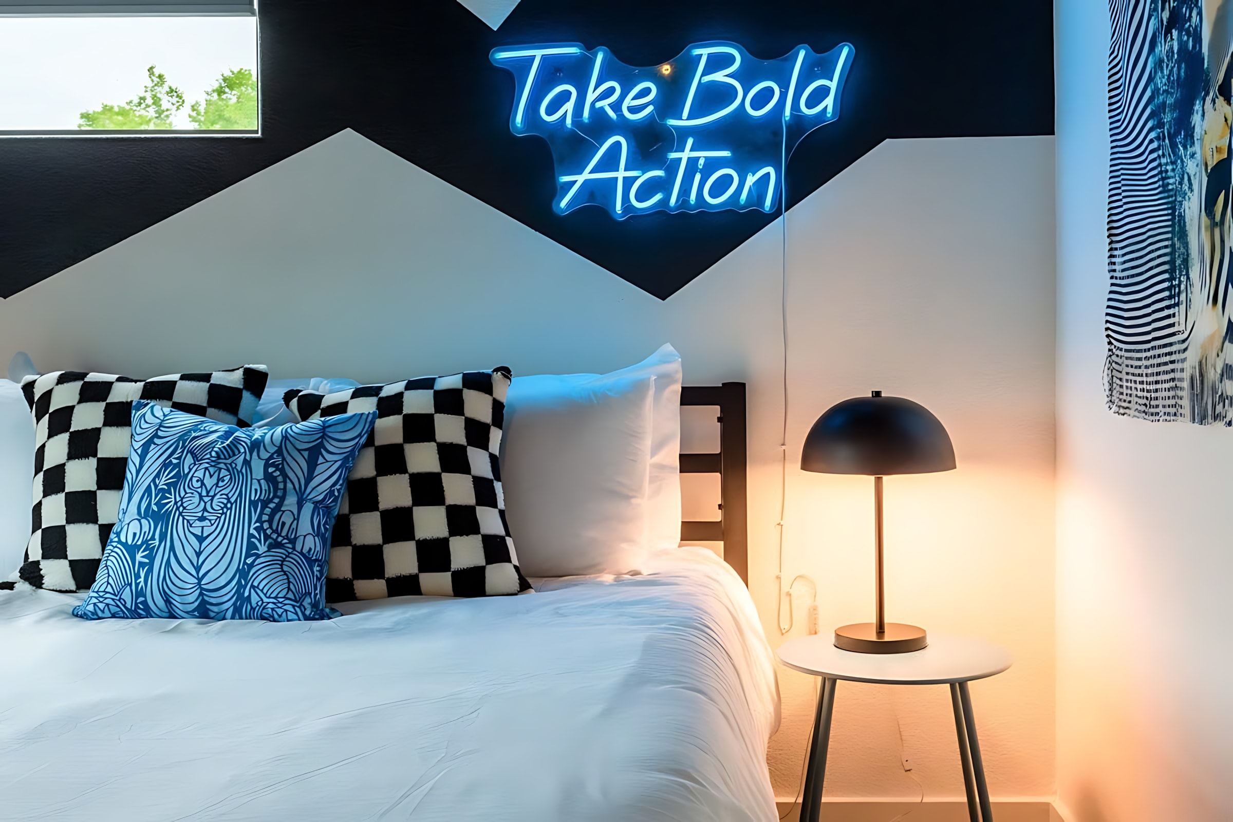 A close-up of a modern bedroom showing a queen bed with white bedding, checkered pillows, and a blue decorative pillow. A bright blue neon sign on the wall reads "Take Bold Action," illuminating the geometric black and white wall design.