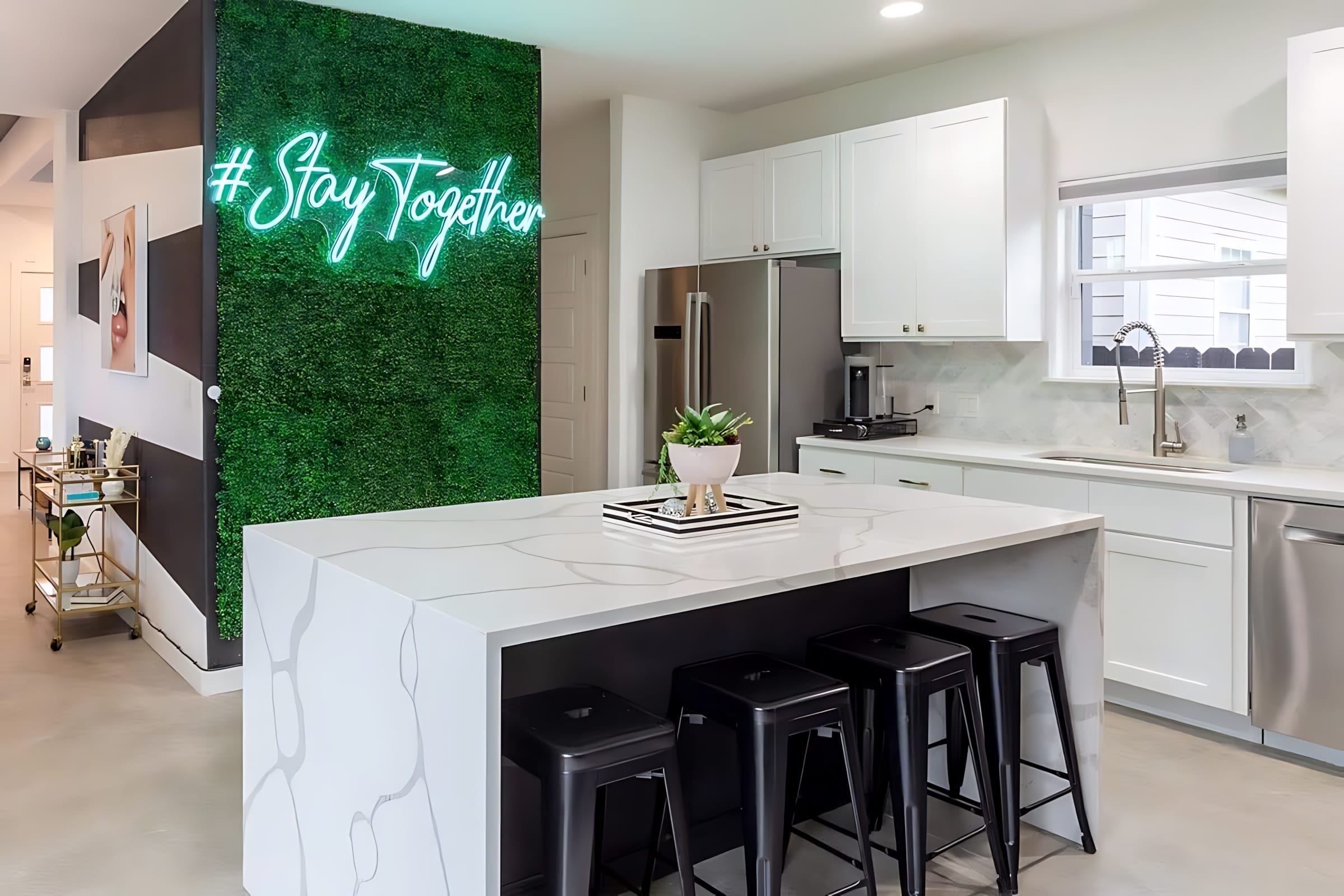 A bright kitchen centered around a large white quartz island with dark base cabinetry and four black industrial-style bar stools, set against a vibrant green faux-plant wall with a glowing neon "#Stay Together" sign. White shaker cabinets and stainless steel appliances line the back wall.