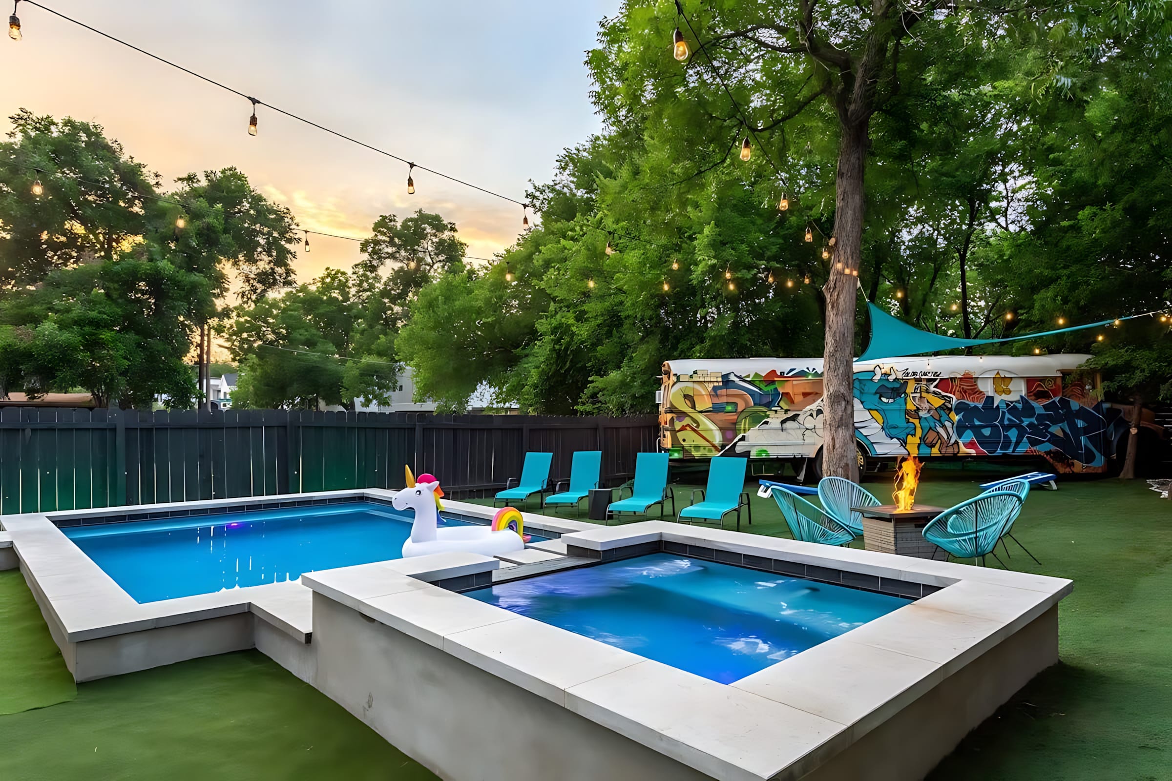 An inviting outdoor space features a rectangular pool with a unicorn float, an adjacent hot tub, and a fire pit surrounded by turquoise chairs, all set on artificial turf under string lights. A colorful, graffiti-covered bus is visible in the background.