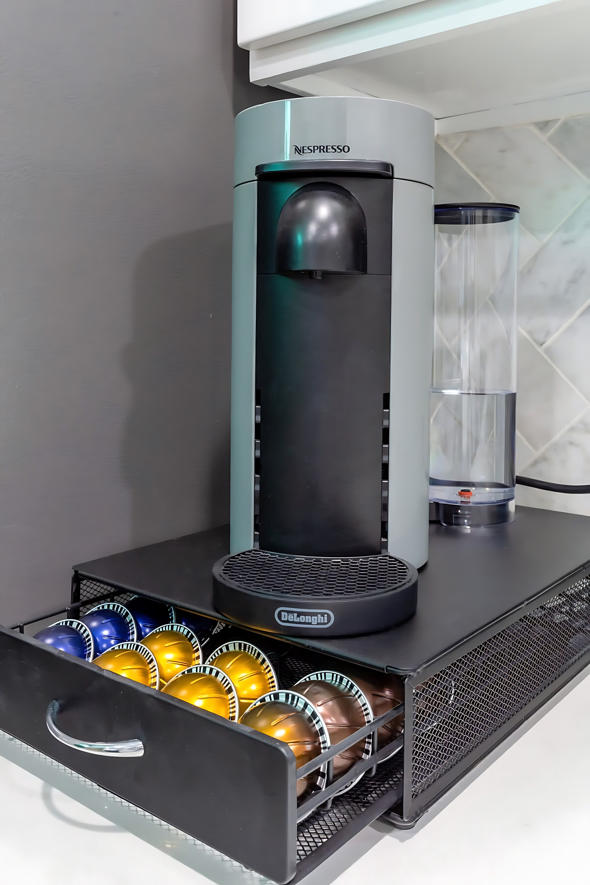 A modern Nespresso coffee machine sits on a black mesh drawer organizer filled with colorful coffee pods, set against a gray wall and white marble-look backsplash.