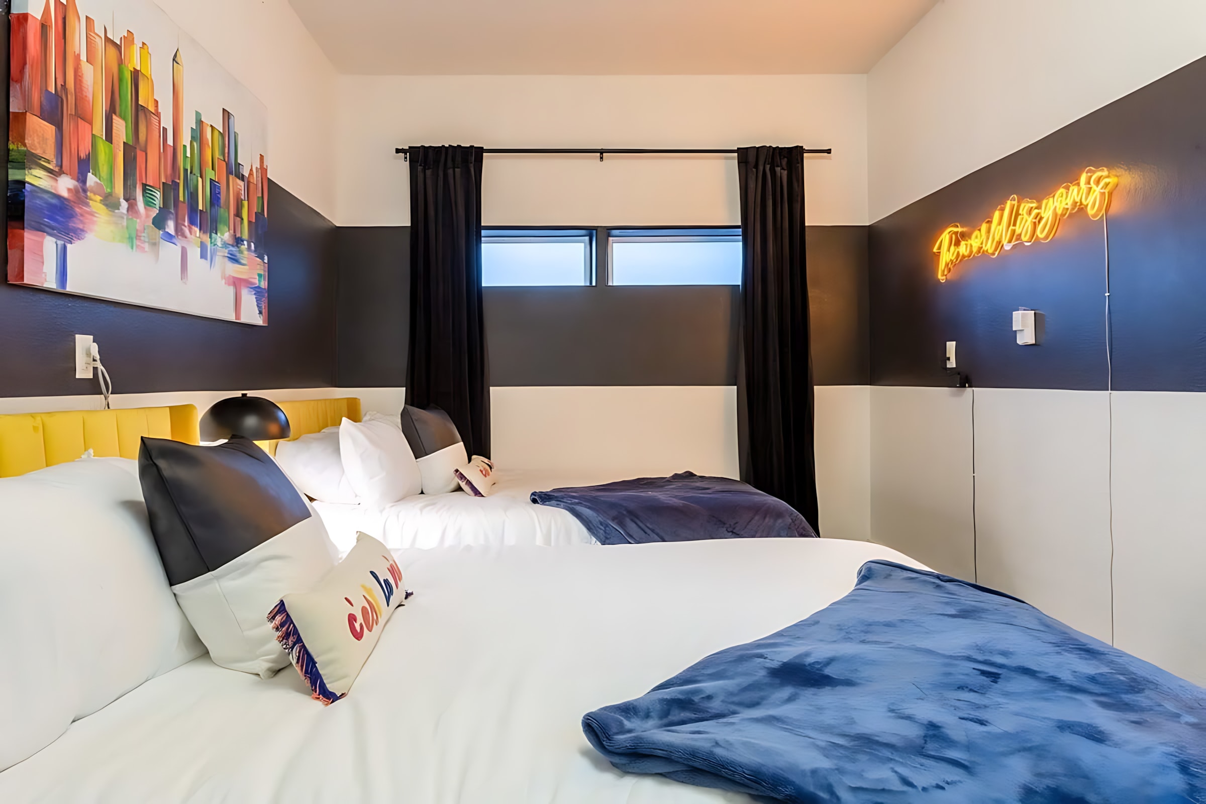 A bedroom with two beds, each with white bedding and blue throws, and a yellow headboard on the left bed. The walls feature a bold black and white horizontal stripe pattern, and a yellow neon sign reads "Treat Yo'self."