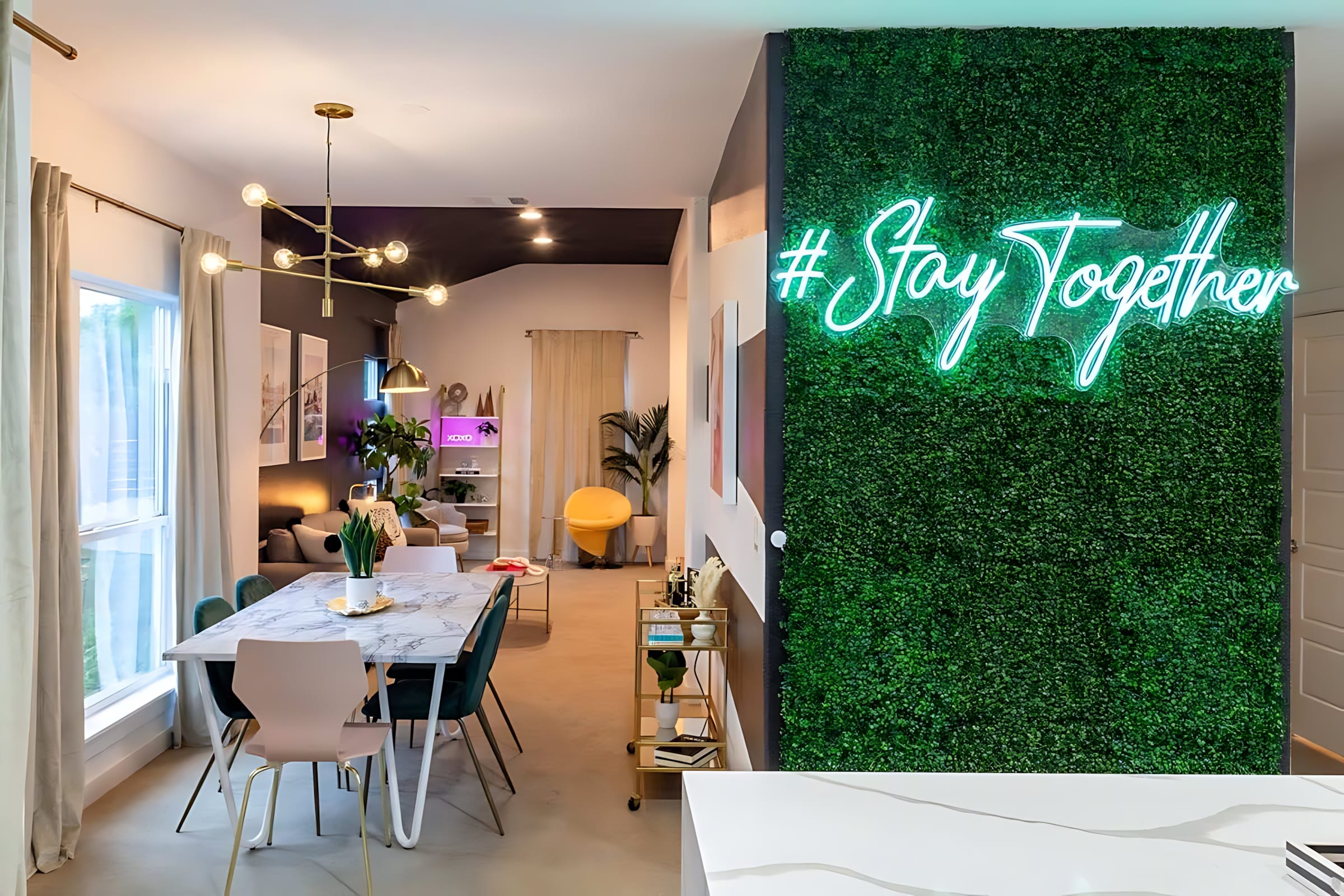 An open-concept dining area with a white marble-top table and mixed-color chairs, illuminated by a modern gold chandelier, leading into a living room. A striking green faux-plant wall with a neon "#Stay Together" sign serves as a vibrant focal point.