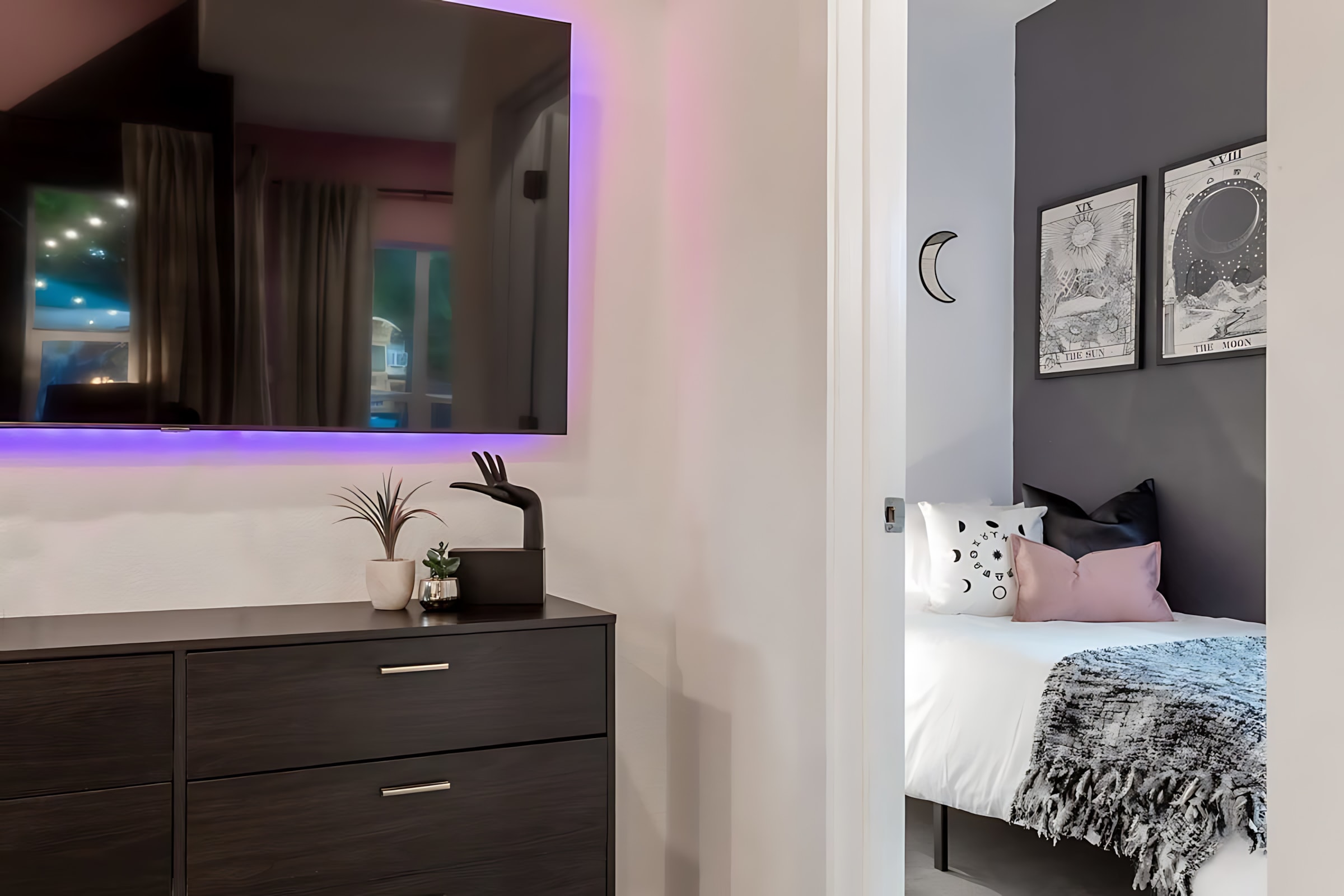 A wall-mounted TV with purple backlighting hangs above a dark wood dresser, with a glimpse into a bedroom featuring moon-themed decor and a single bed through an open doorway.