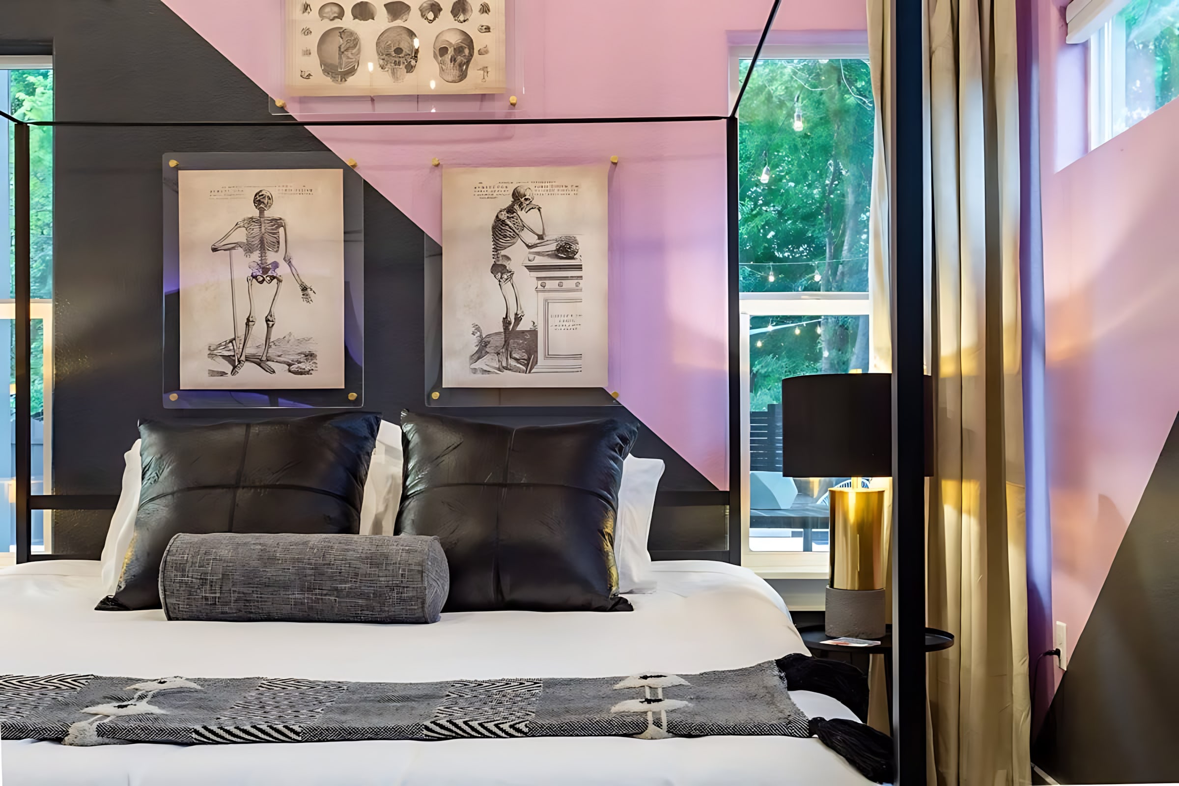 A modern bedroom features a large bed with black leather pillows and a gray bolster, set against a striking black and pink geometric accent wall adorned with framed anatomical prints.