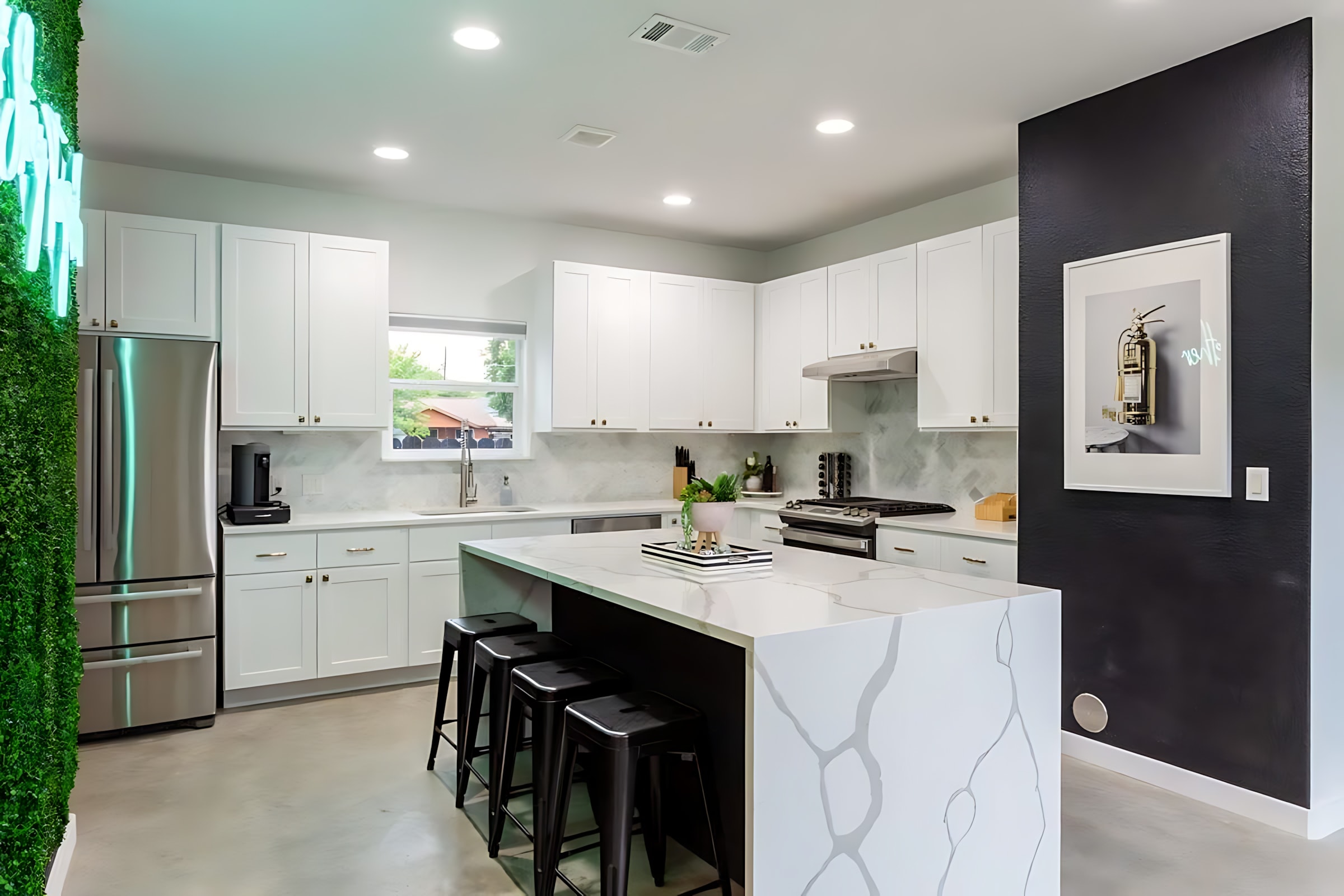 A spacious, modern kitchen featuring white cabinetry, stainless steel appliances including a large refrigerator, and a substantial white quartz island with seating for four black bar stools. A green faux-plant wall with a neon sign is visible on the left.