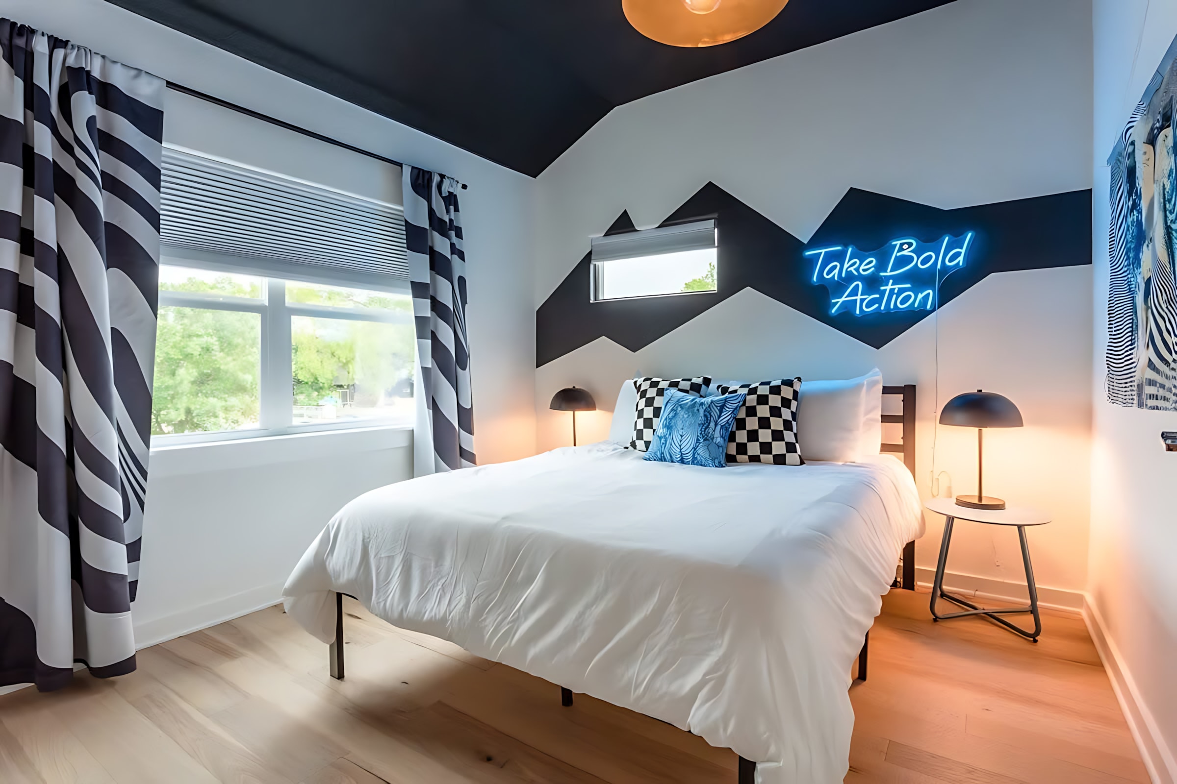 A modern bedroom with a queen-sized bed, black and white striped curtains, and a striking blue neon sign on the wall that reads "Take Bold Action." The walls feature a geometric black and white pattern.