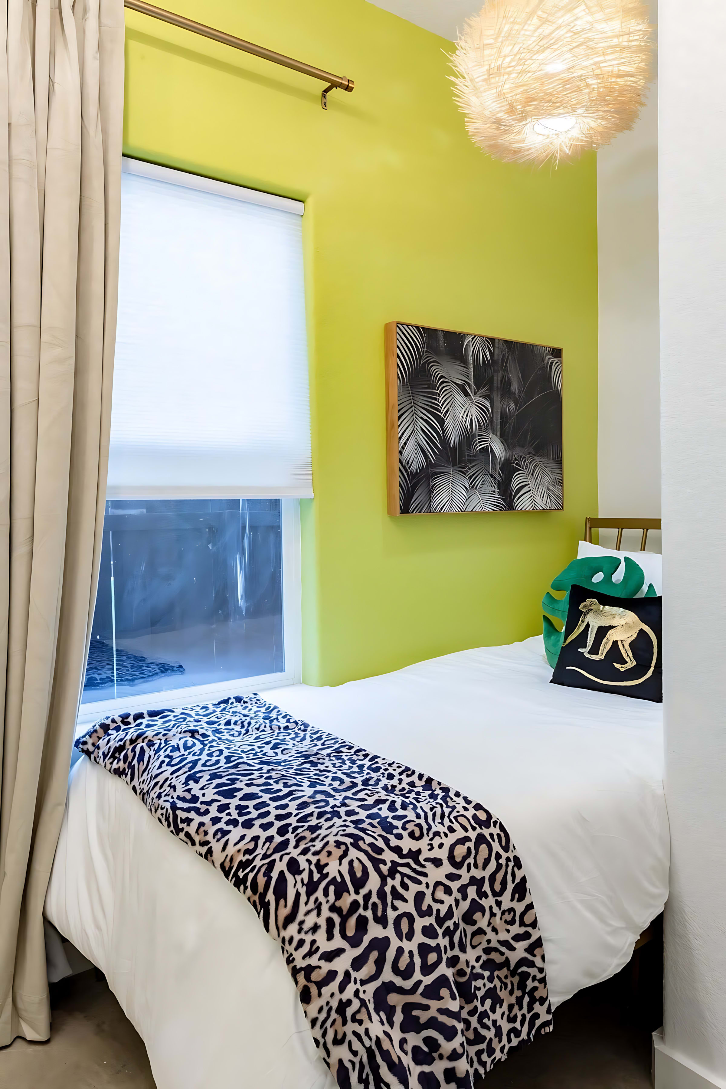 A cozy bedroom featuring a single bed with a leopard print throw blanket and a lime green accent wall. A framed black and white botanical print hangs above the bed, and a unique woven light fixture illuminates the space.