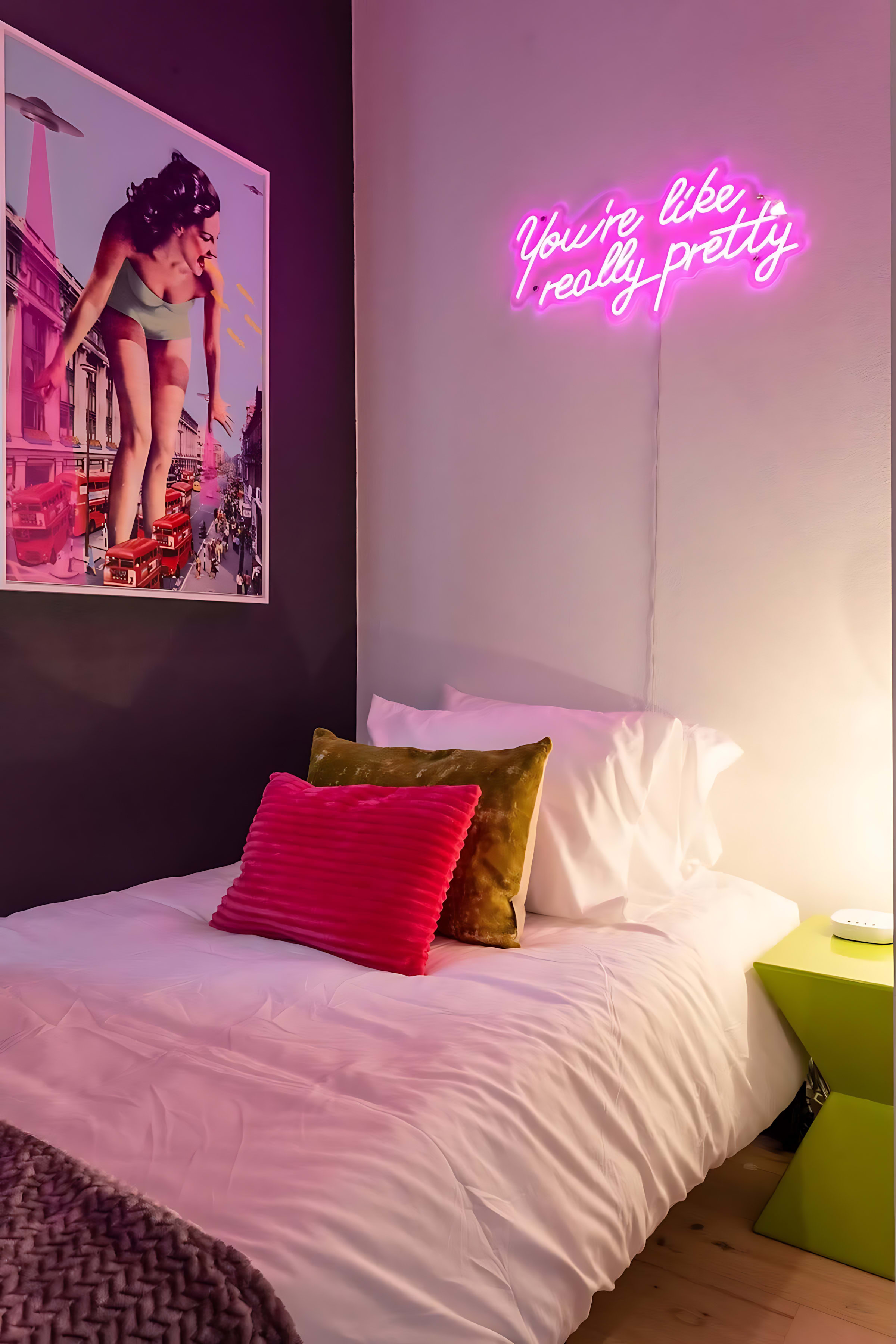 A close-up of a vibrant small bedroom with a single bed, a "You're like, really pretty" neon sign, and a large pop art print on the wall, complemented by colorful throw pillows.