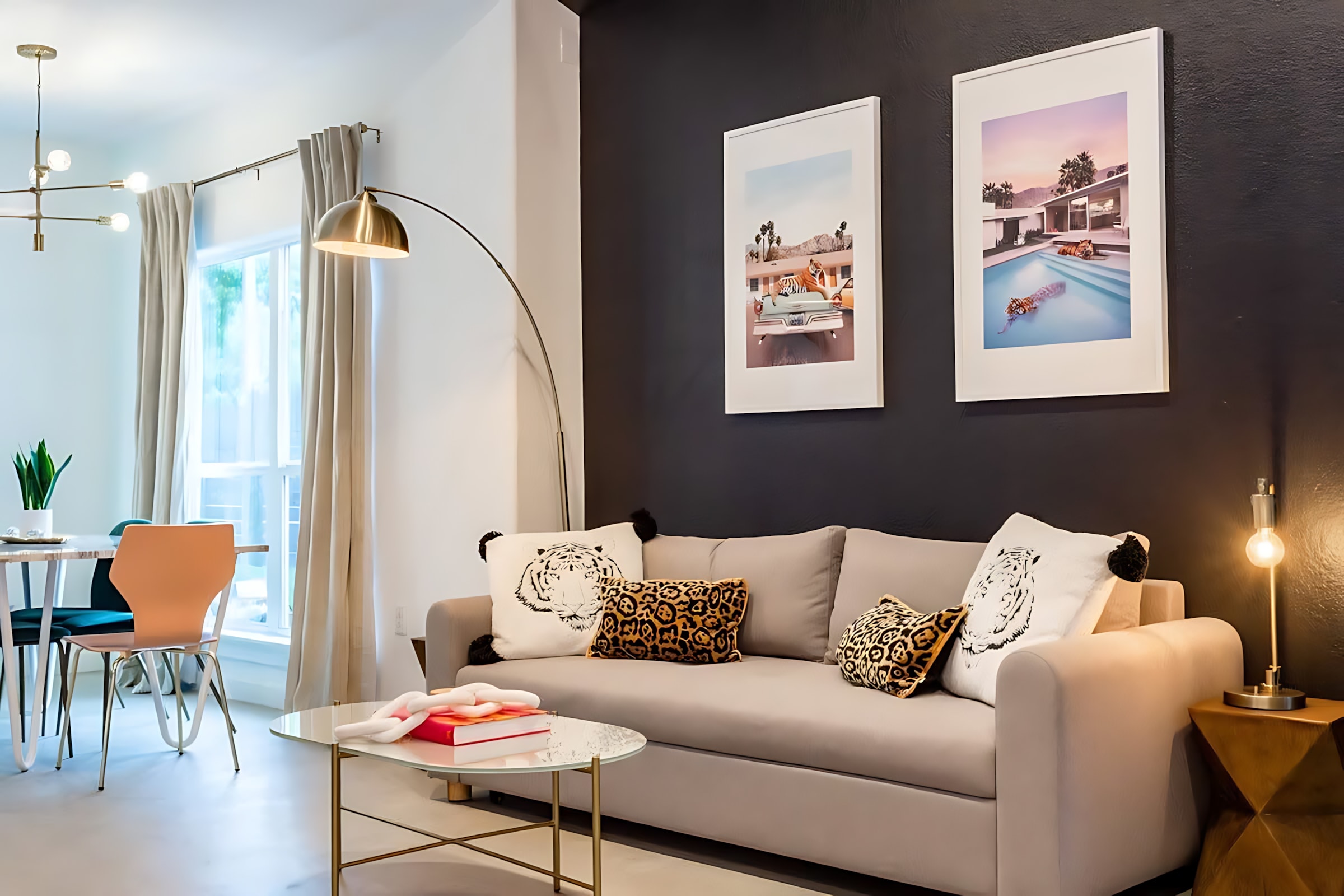 A modern living room featuring a light gray sofa with decorative pillows, a glass coffee table, and a gold arc floor lamp, set against a dark accent wall with two framed art prints. A dining area with a white table and colorful chairs is visible in the background.