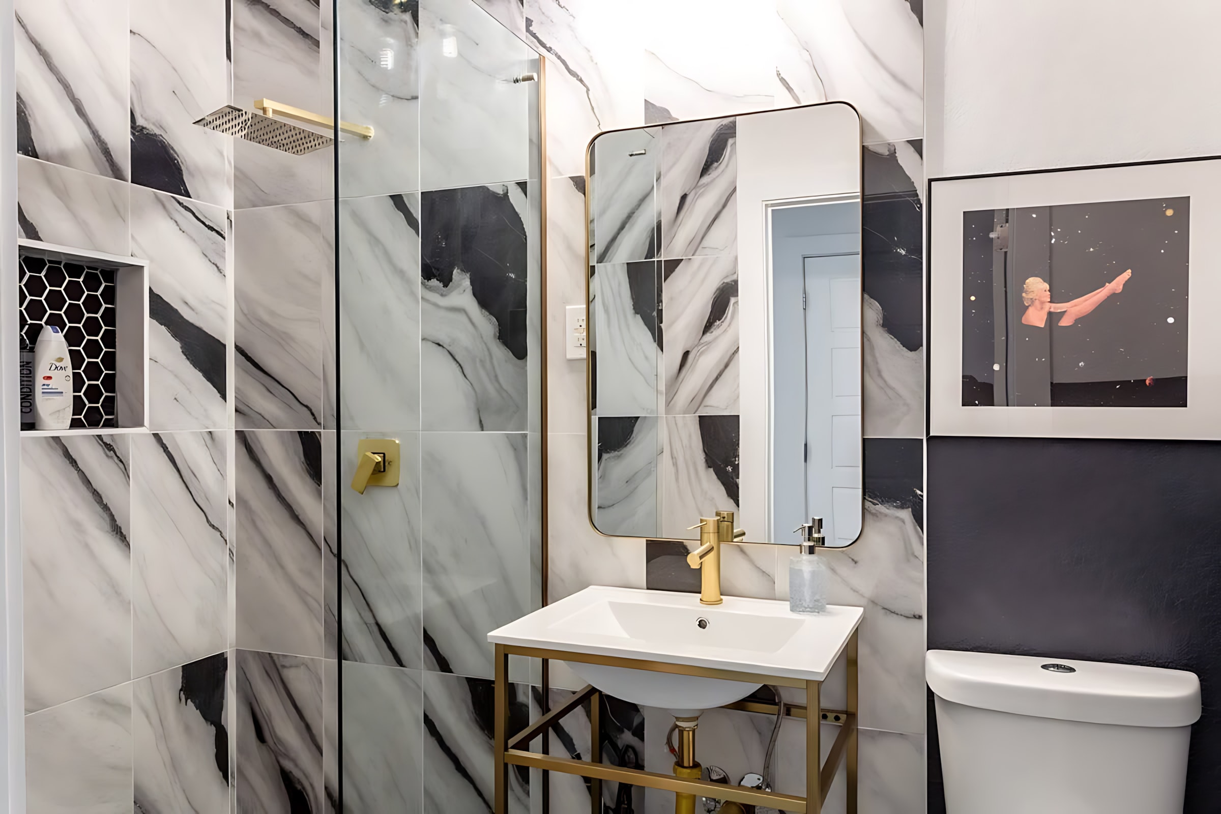 A modern bathroom featuring black and white marble-patterned tiles, a gold-framed rectangular mirror, and gold-finished faucet and shower fixtures. A built-in shower niche with hexagonal black tiles holds toiletries.