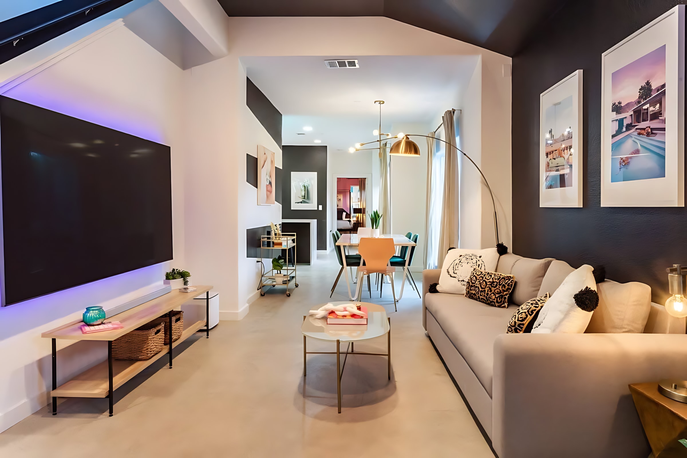 An open-concept living space with a large wall-mounted TV and entertainment console on the left, leading to a light gray sofa and glass coffee table on the right, with a dining area visible further back. The room features a mix of white and dark accent walls and modern lighting.