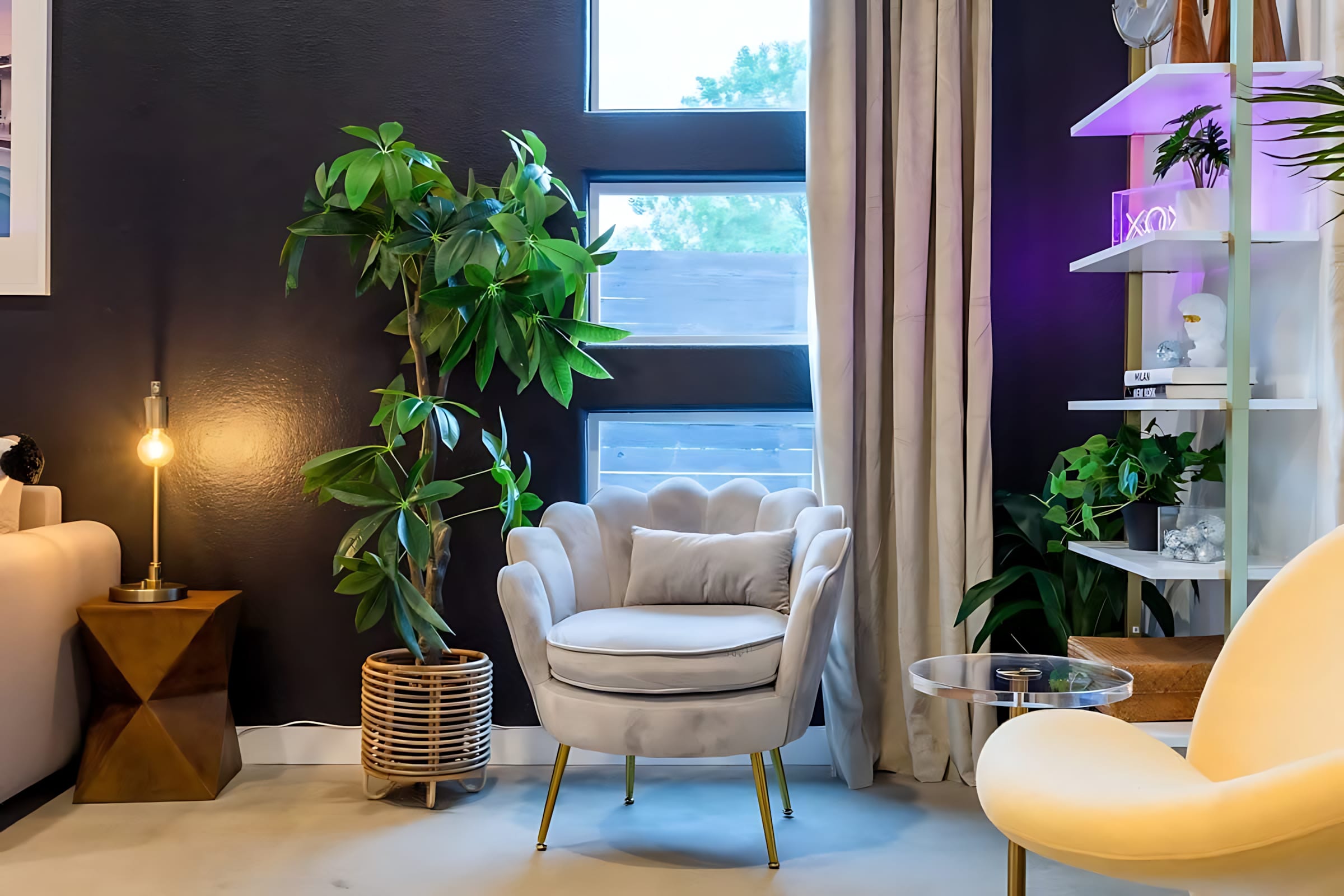A cozy corner of the living room featuring a plush, shell-back armchair with gold legs, positioned next to a tall potted plant in a woven basket and a modern table lamp. Large windows provide natural light, and a white shelving unit with decorative items is visible in the background.