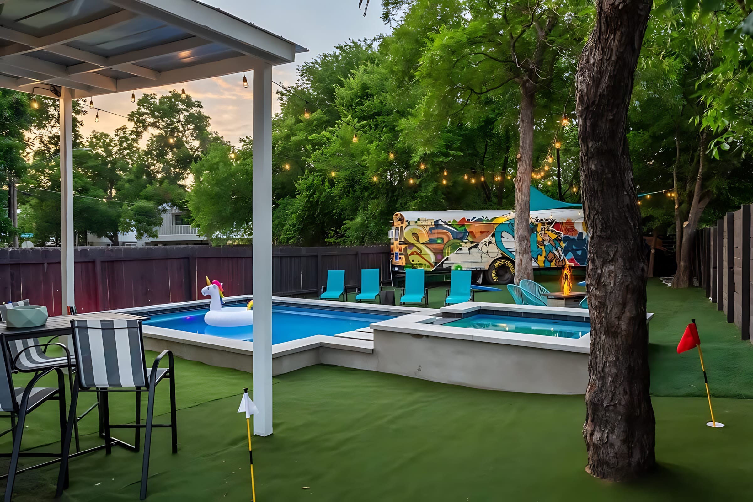 A lively backyard features a rectangular pool with a unicorn float, an adjacent hot tub, and a putting green with flags. A colorful, graffiti-covered school bus is parked in the background, surrounded by lush trees and string lights.