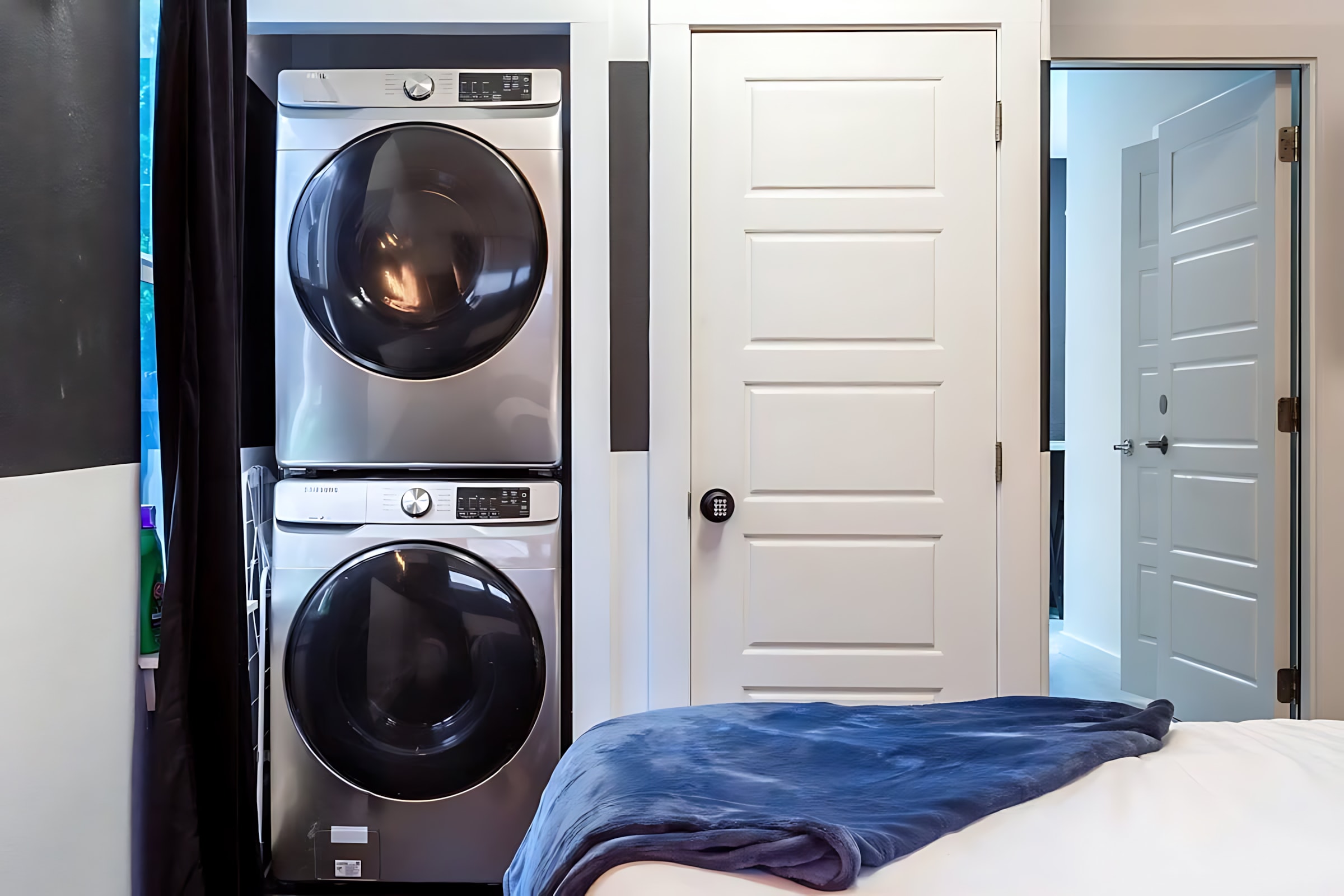 A stacked silver washer and dryer unit is neatly tucked into an alcove next to a white paneled door, with a blue throw blanket visible on a bed in the foreground.