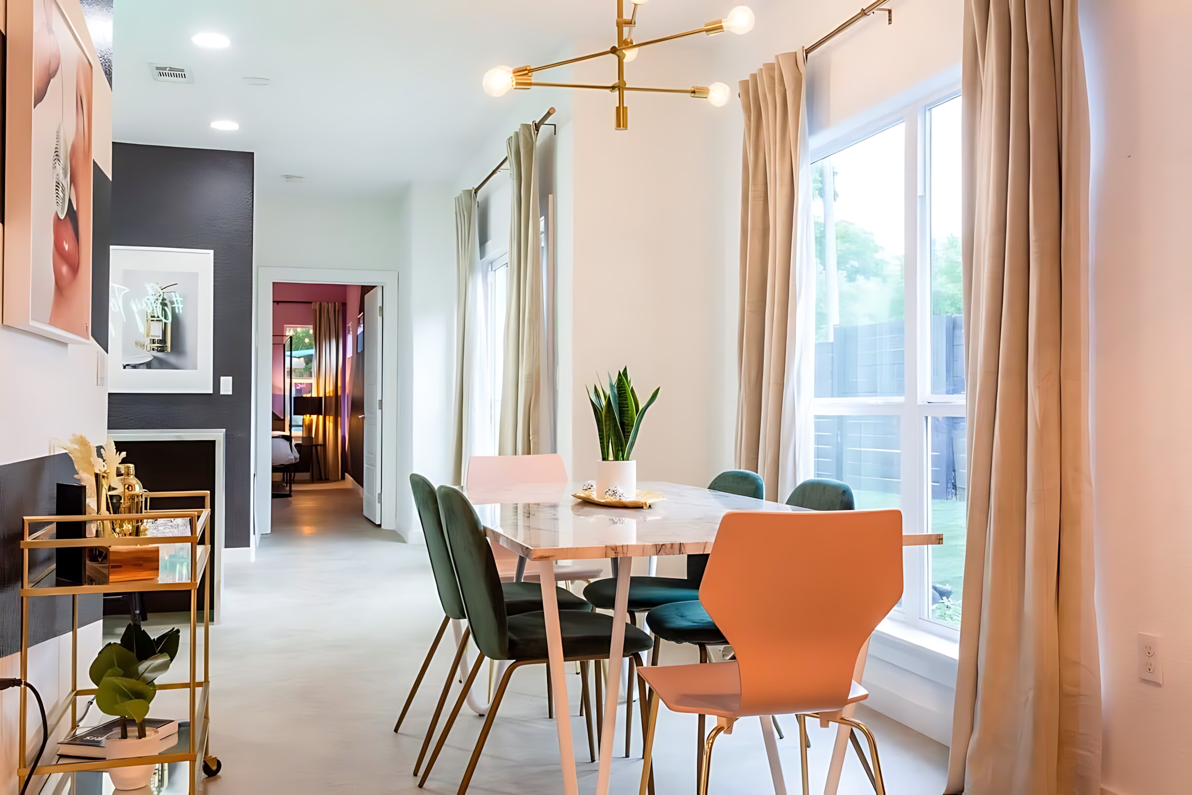 A bright dining area featuring a white marble-top table surrounded by four dark green velvet chairs and two peach-colored chairs, beneath a modern gold chandelier. Large windows with light drapes provide ample natural light, and a gold bar cart is visible to the left.