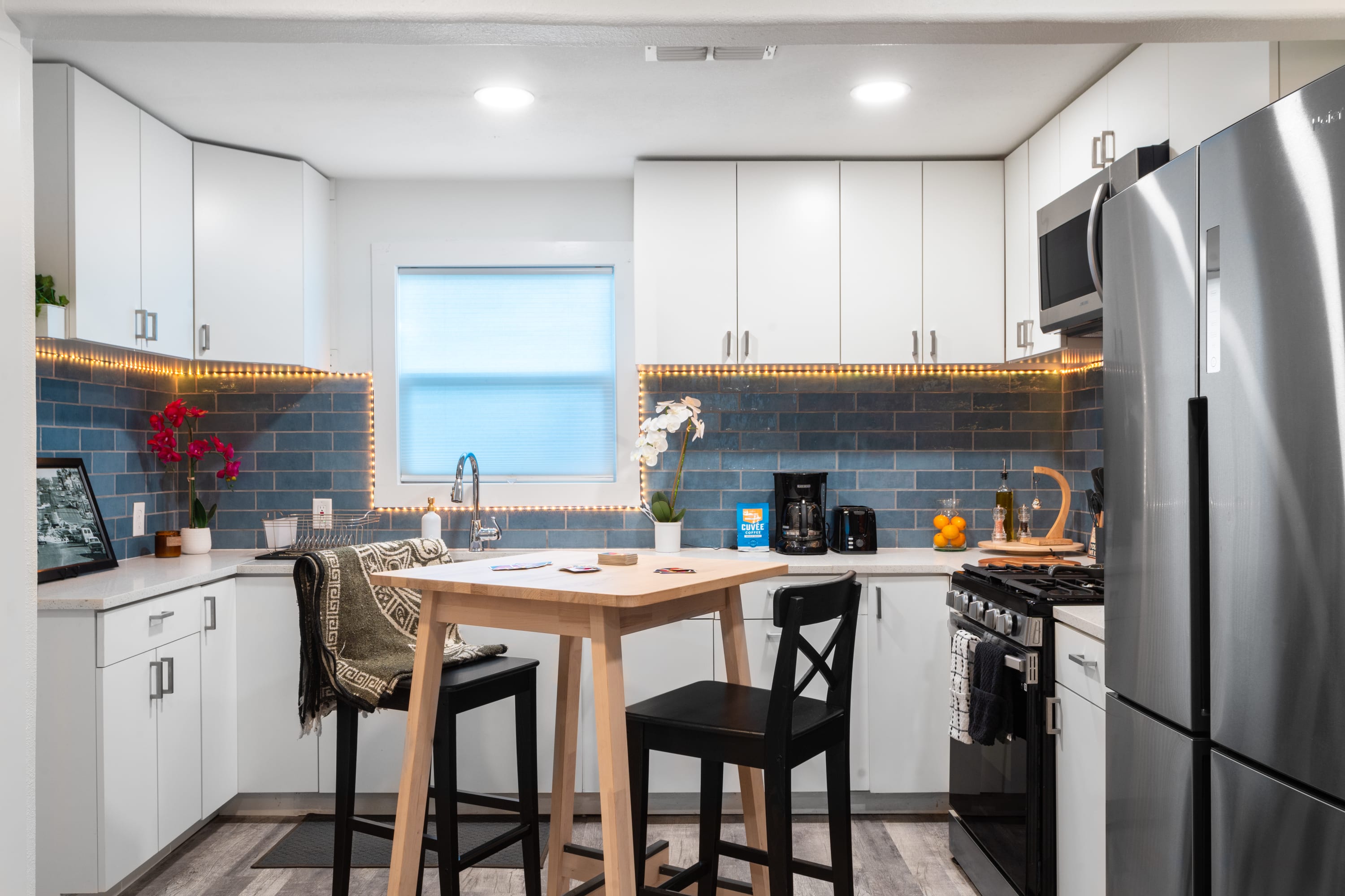 A wide shot of the modern kitchen, showcasing white shaker cabinets, light countertops, and a blue subway tile backsplash with under-cabinet LED lighting. A wooden high-top table with two black stools is centrally placed, and stainless steel appliances are prominent.