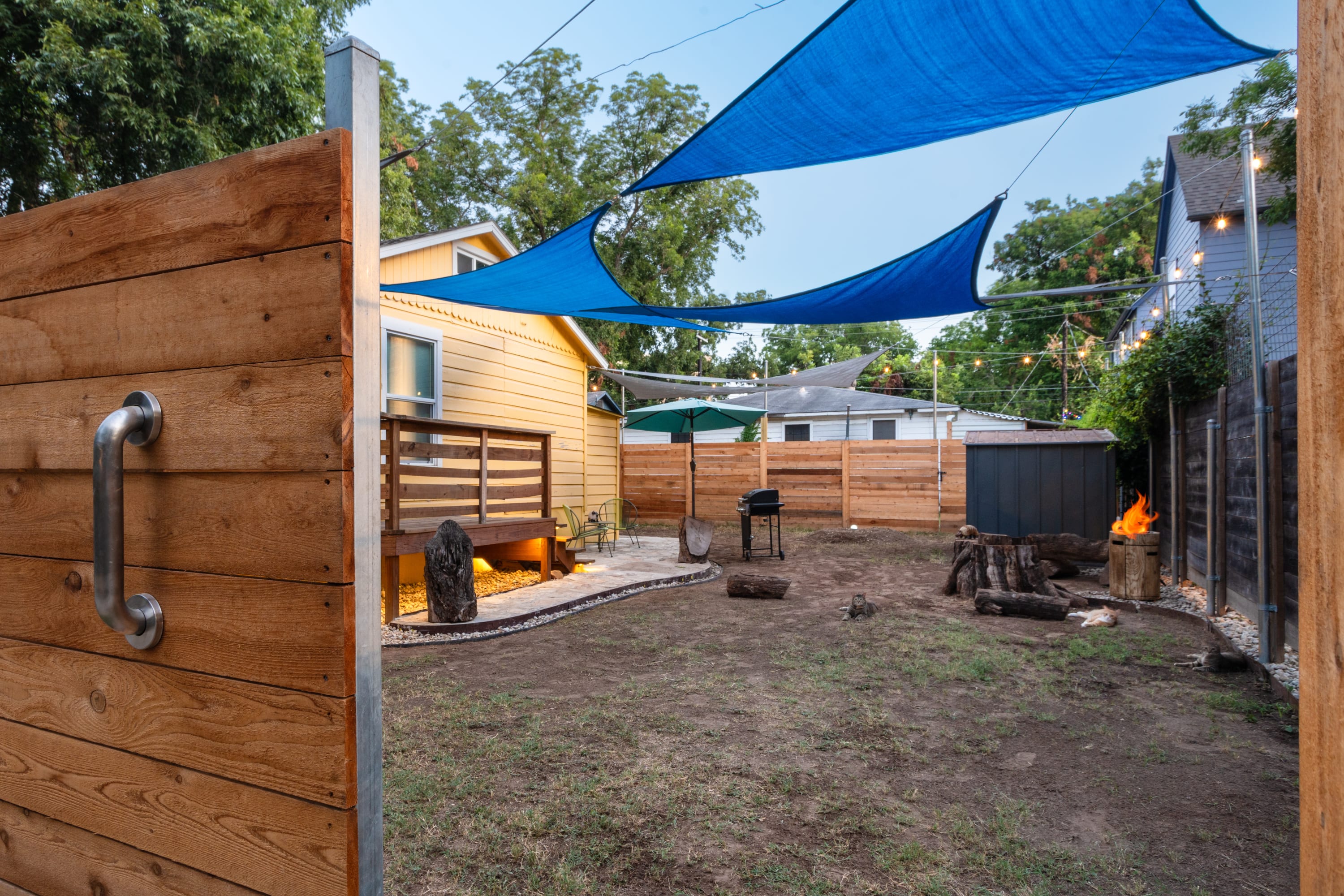 Spacious backyard with a fire pit, a grill, and a paved patio area leading to a yellow house with a wooden deck. Blue and grey shade sails are strung overhead, and a wooden fence surrounds the property.