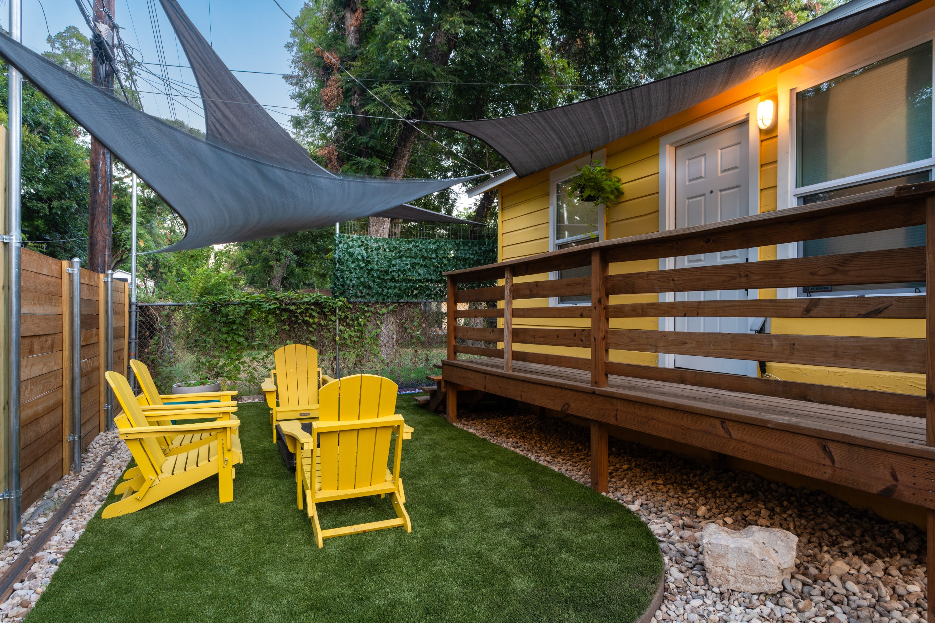 Inviting outdoor seating area with four bright yellow Adirondack chairs arranged around a rustic wooden coffee table on a patch of artificial turf. Grey shade sails provide cover, and a wooden fence encloses the space.