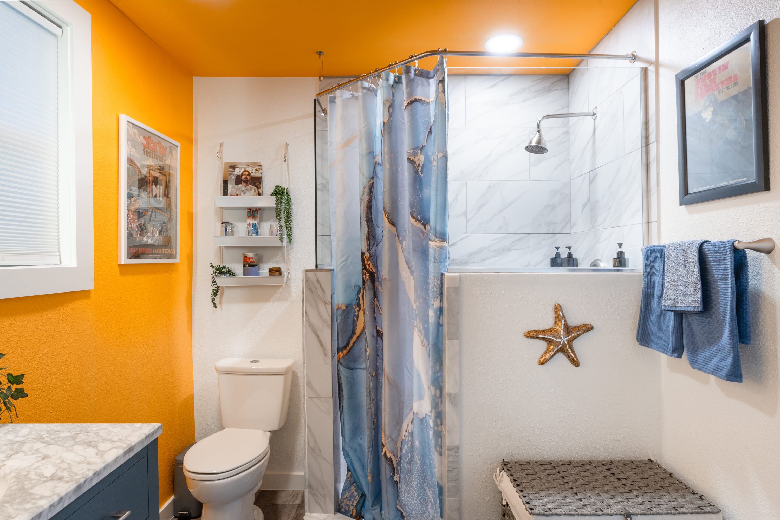Modern bathroom featuring a walk-in shower with white subway tiles and a blue and gold marble-patterned shower curtain. The walls and ceiling are bright orange, and a white toilet is positioned next to a window.