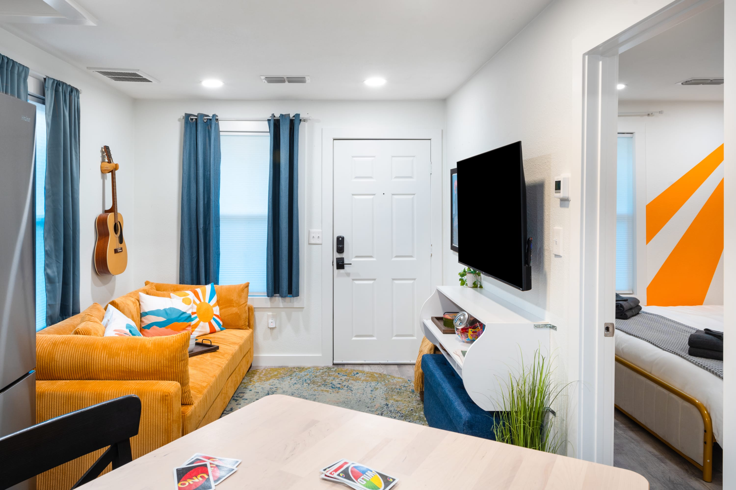 A view of the living room from the kitchen, showing a mustard yellow corduroy sofa with colorful pillows, a wall-mounted TV, and a white front door. The kitchen's wooden high-top table is partially visible in the foreground.