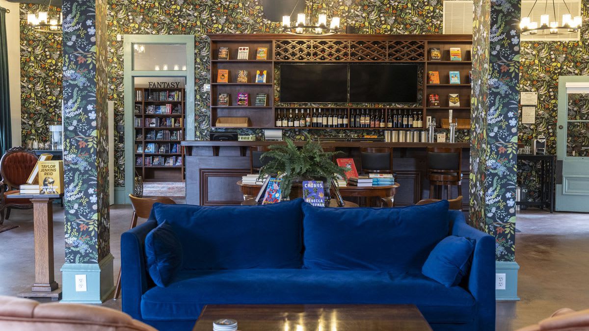 A vibrant common area features a deep blue velvet sofa in the foreground, facing a wooden coffee table. The walls are adorned with a busy floral wallpaper, and a bar area with bookshelves and a TV is visible in the background.