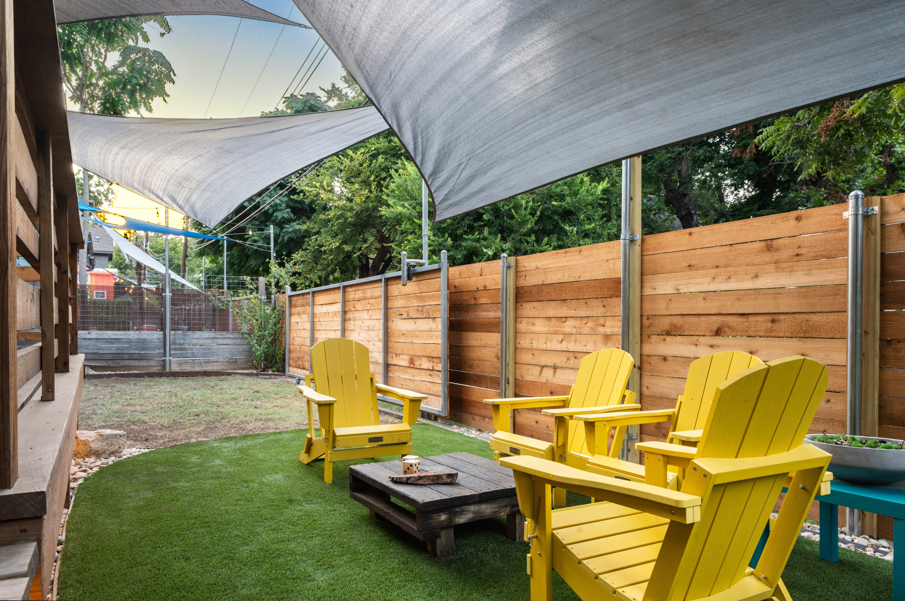 Expansive backyard area featuring a section of artificial turf with yellow Adirondack chairs and a wooden coffee table, shaded by large grey sails. A tall wooden fence provides privacy around the perimeter.