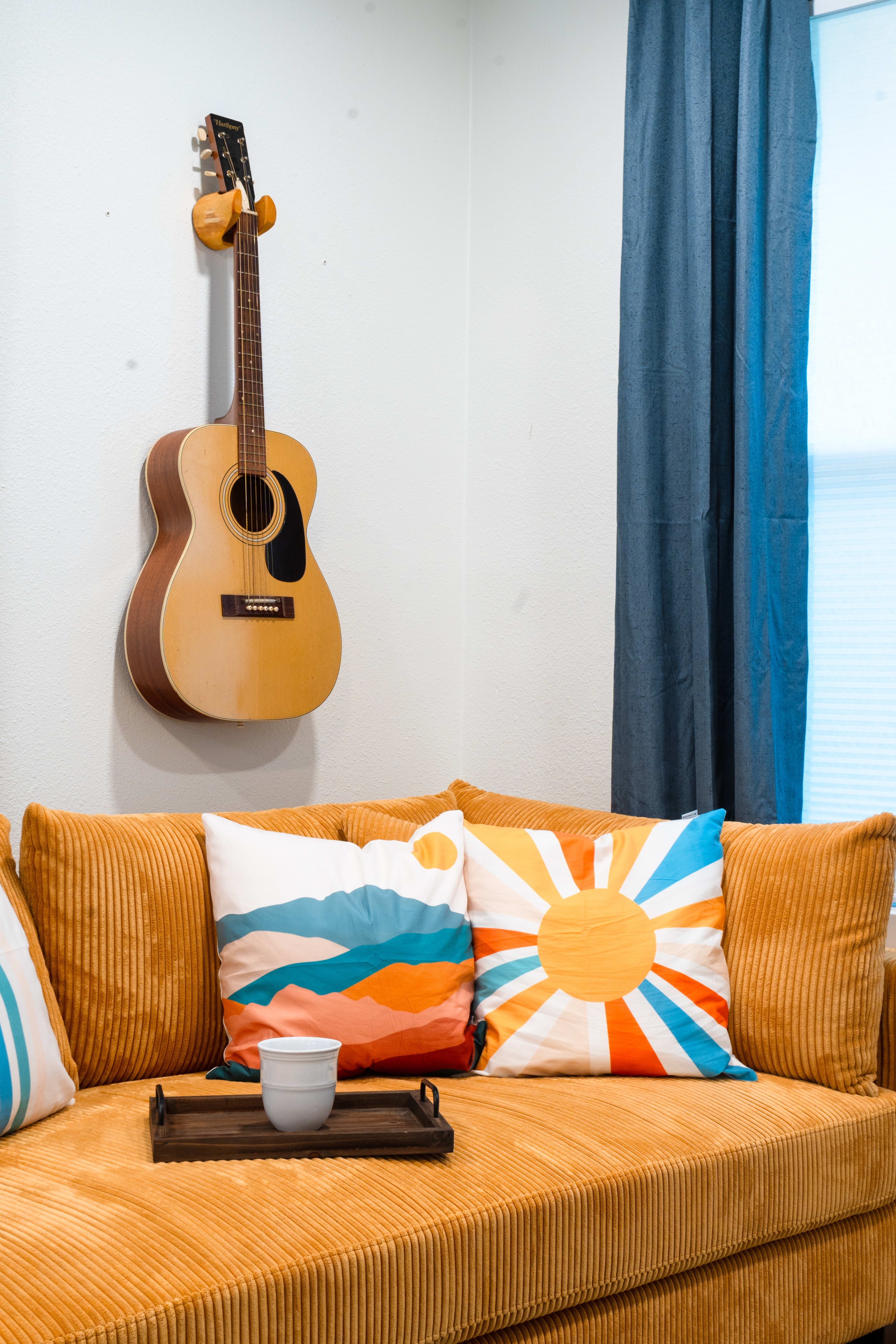 A close-up of the mustard yellow corduroy sofa in the living room, adorned with vibrant throw pillows featuring abstract and sunburst designs. An acoustic guitar hangs on the white wall above the sofa, next to blue curtains.