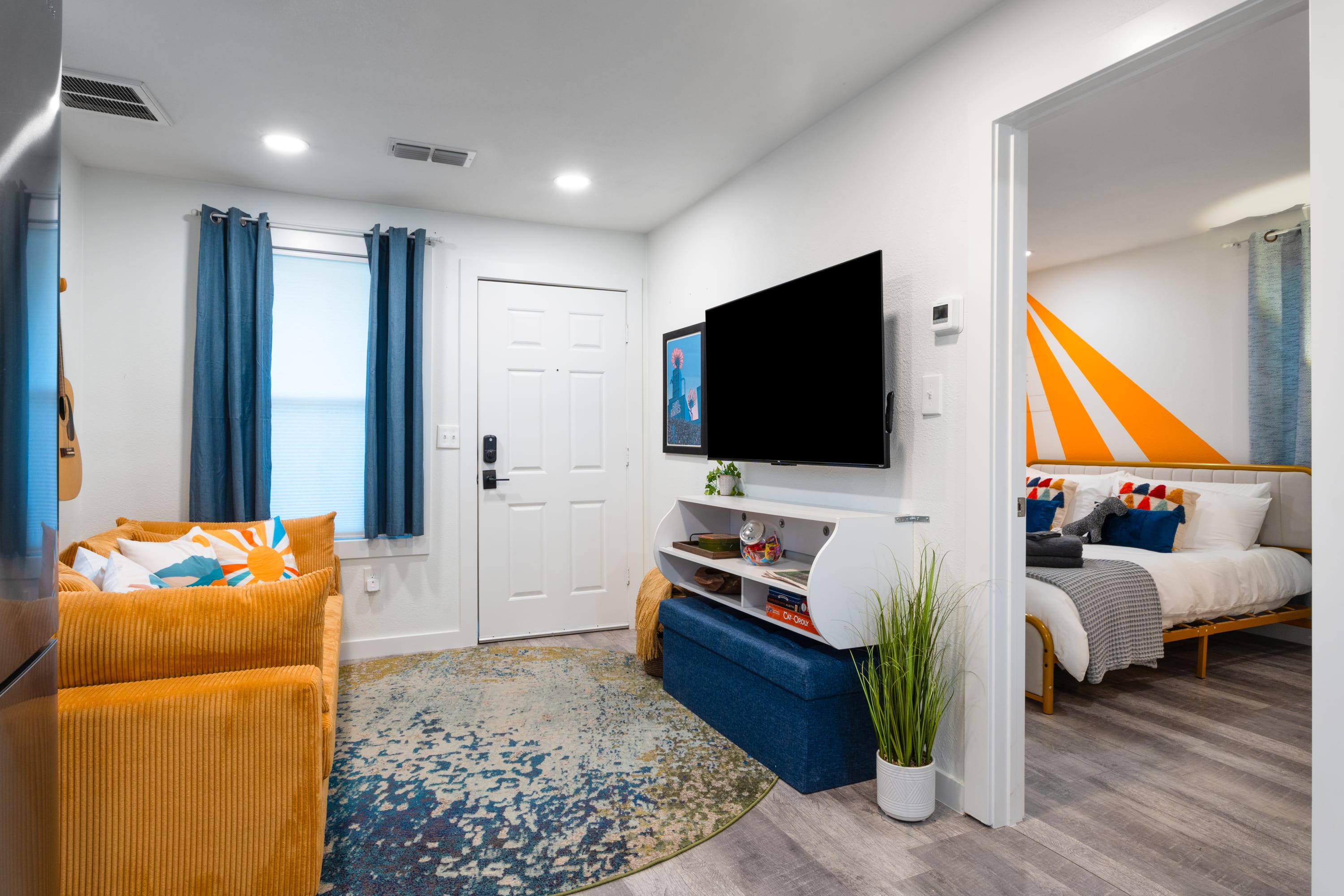 Bright living room featuring a mustard yellow corduroy sofa, a wall-mounted TV above a white media console, and a colorful abstract rug on light wood-look flooring. A doorway leads to a bedroom with an orange accent wall.