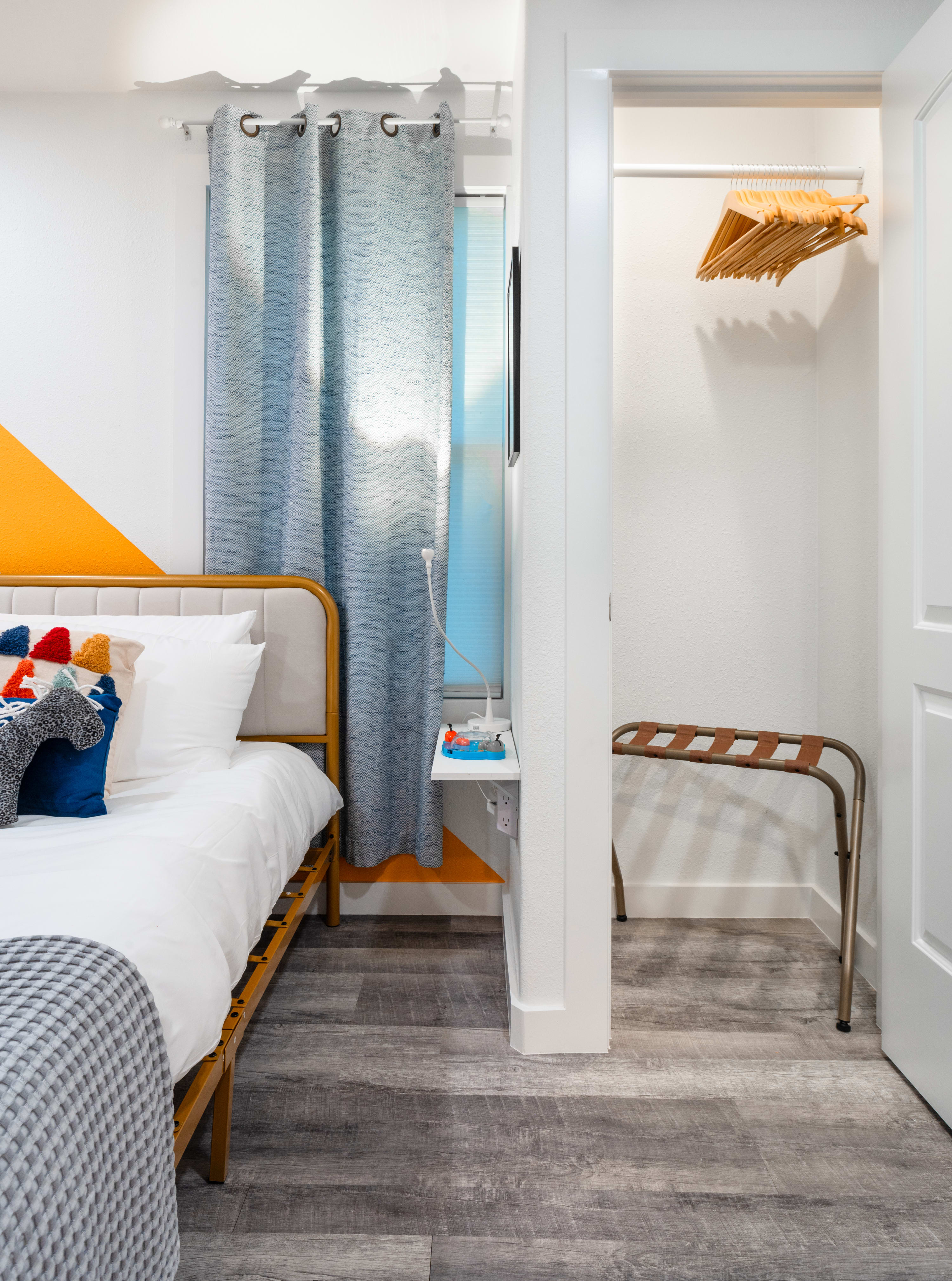 Cozy bedroom with a gold-framed bed featuring white bedding and colorful throw pillows, an orange geometric accent wall, and a window with light blue curtains. A small open closet with wooden hangers is visible to the right.