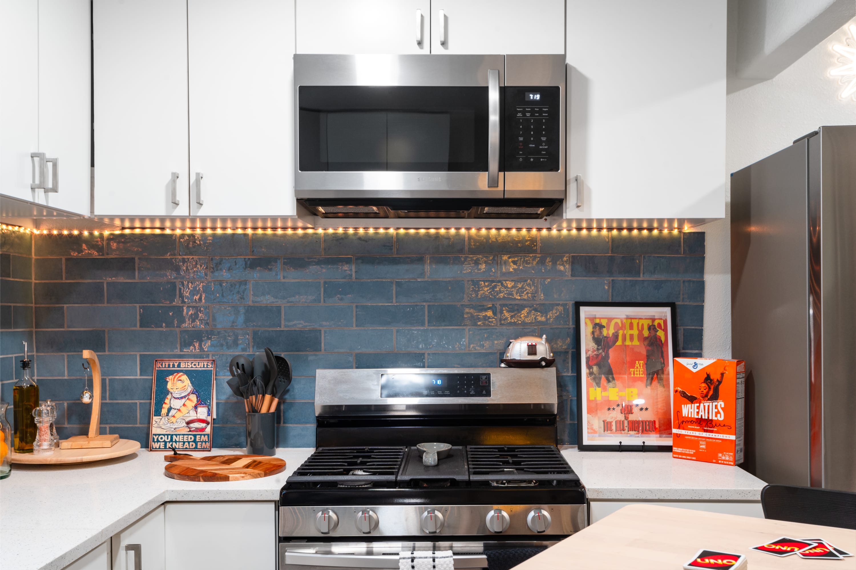 A detailed view of the kitchen's cooking area, featuring a stainless steel gas range with an overhead microwave, set against a blue subway tile backsplash with warm LED strip lighting. White countertops display various kitchen items and decorative elements.
