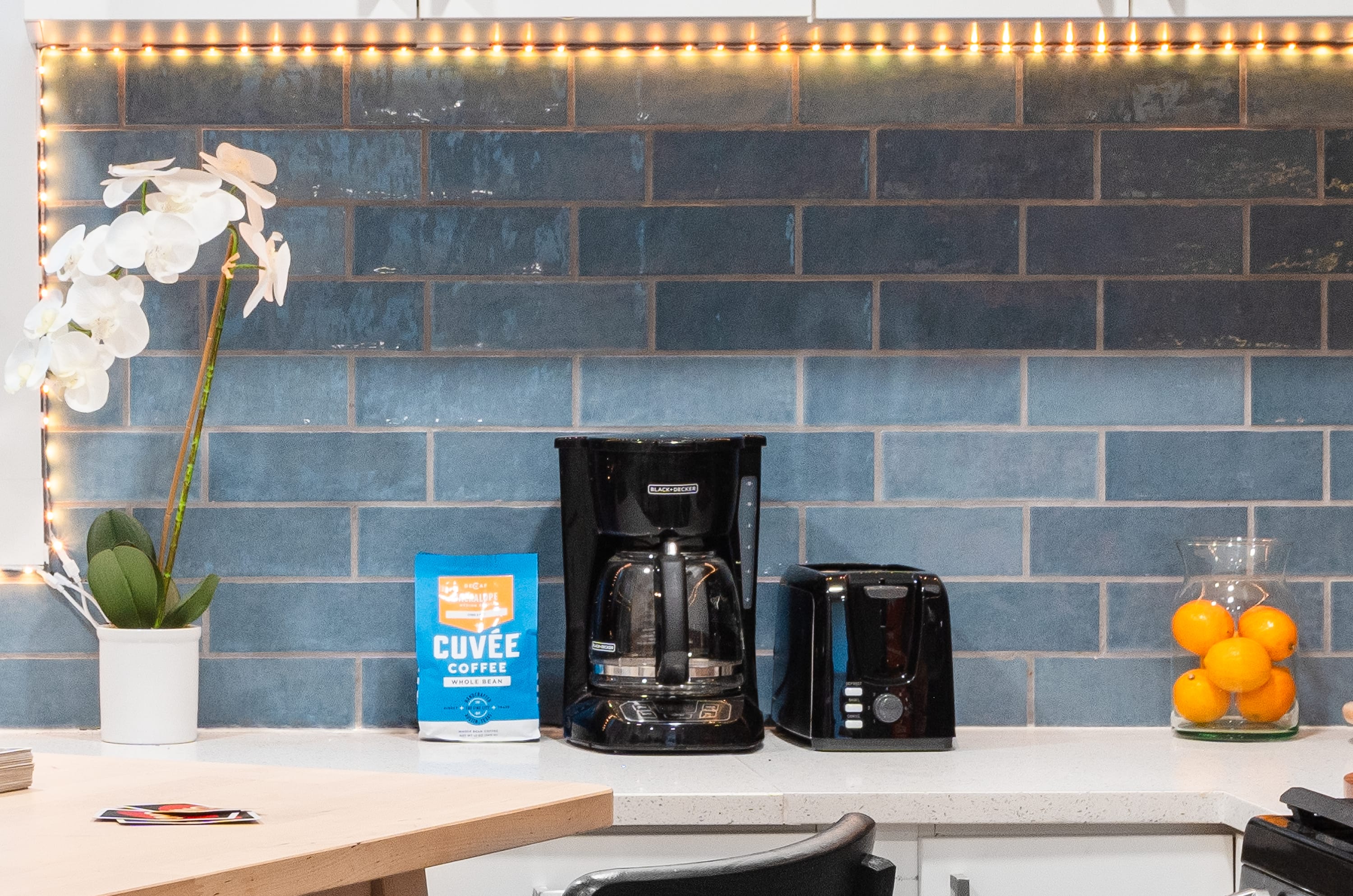 A close-up of the kitchen counter, highlighting a coffee maker, toaster, and a bag of coffee beans, positioned next to a white orchid plant. The blue subway tile backsplash and warm LED strip lighting create an inviting atmosphere.