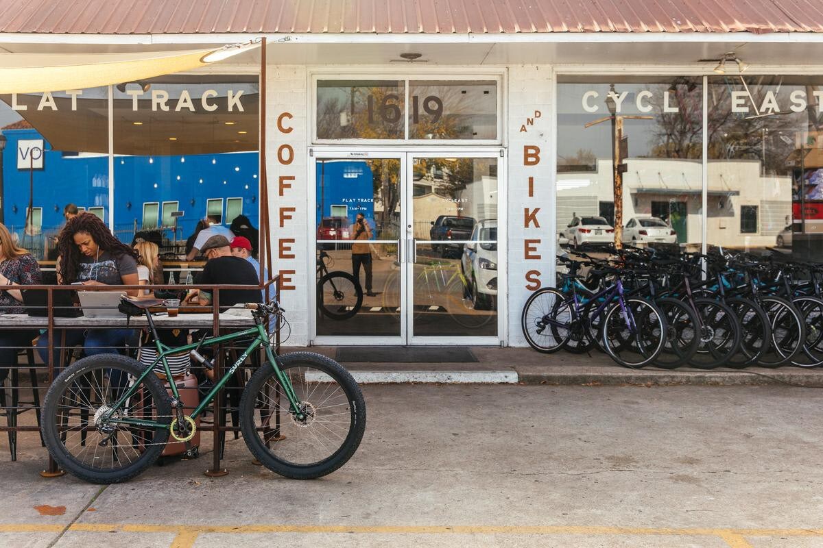 The exterior of 'Flat Track Coffee and Bikes' features a large storefront with glass doors and windows, displaying bicycles for sale. Customers are seated at outdoor tables under an awning, enjoying coffee.