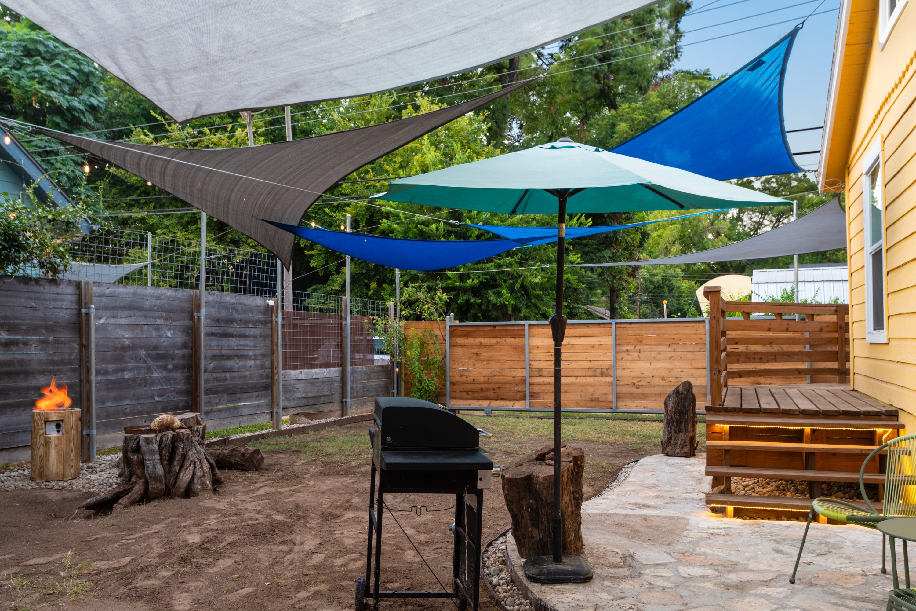 Outdoor entertainment area in the backyard featuring a black gas grill, a fire pit with burning logs, and a teal umbrella providing shade. The area is partially paved with stone and surrounded by dirt and wood stumps.