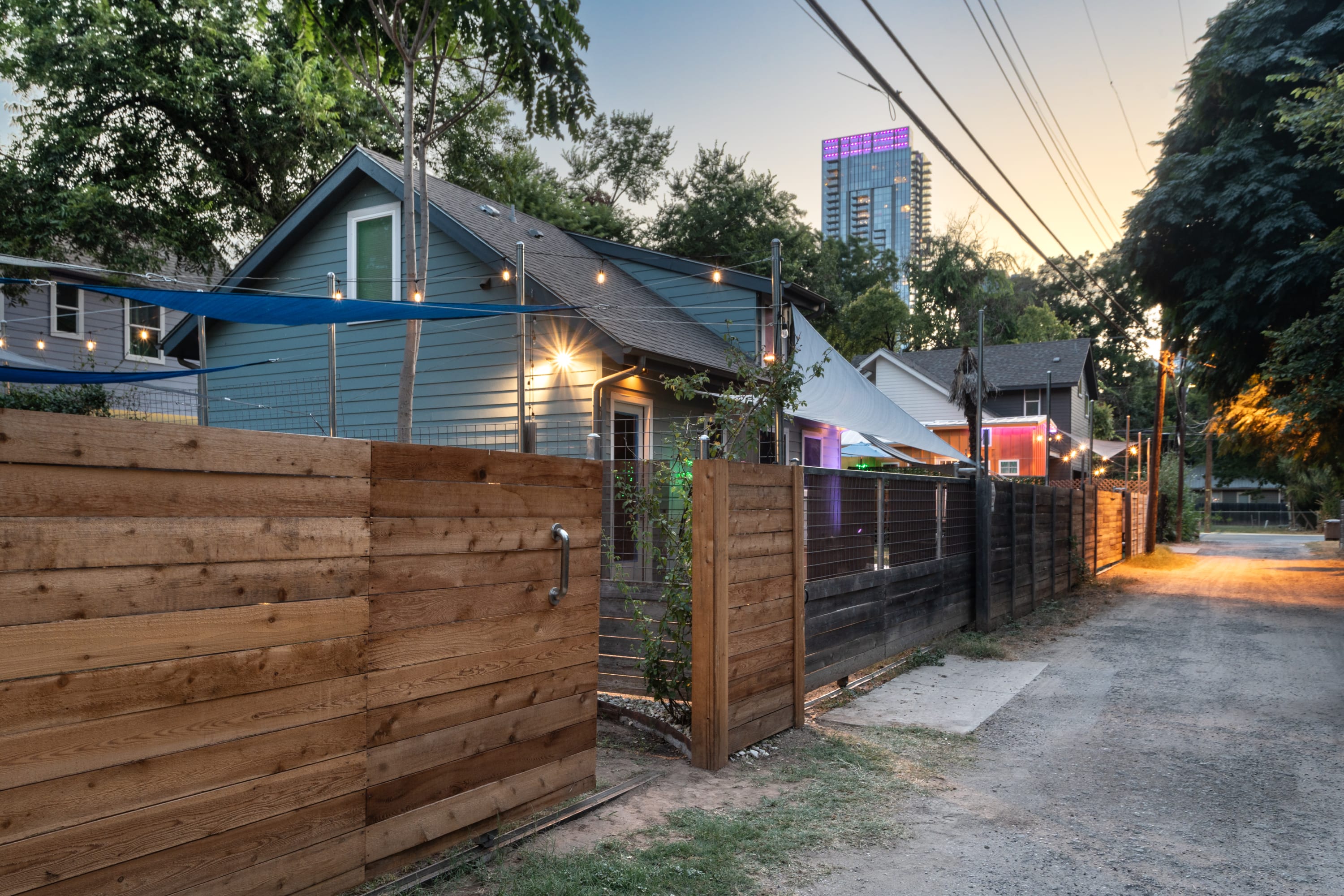 The light blue bungalow is seen from an alleyway at dusk, with string lights illuminating the yard and a tall modern building in the distance. A wooden fence separates the property from the gravel alley, which has power lines overhead.