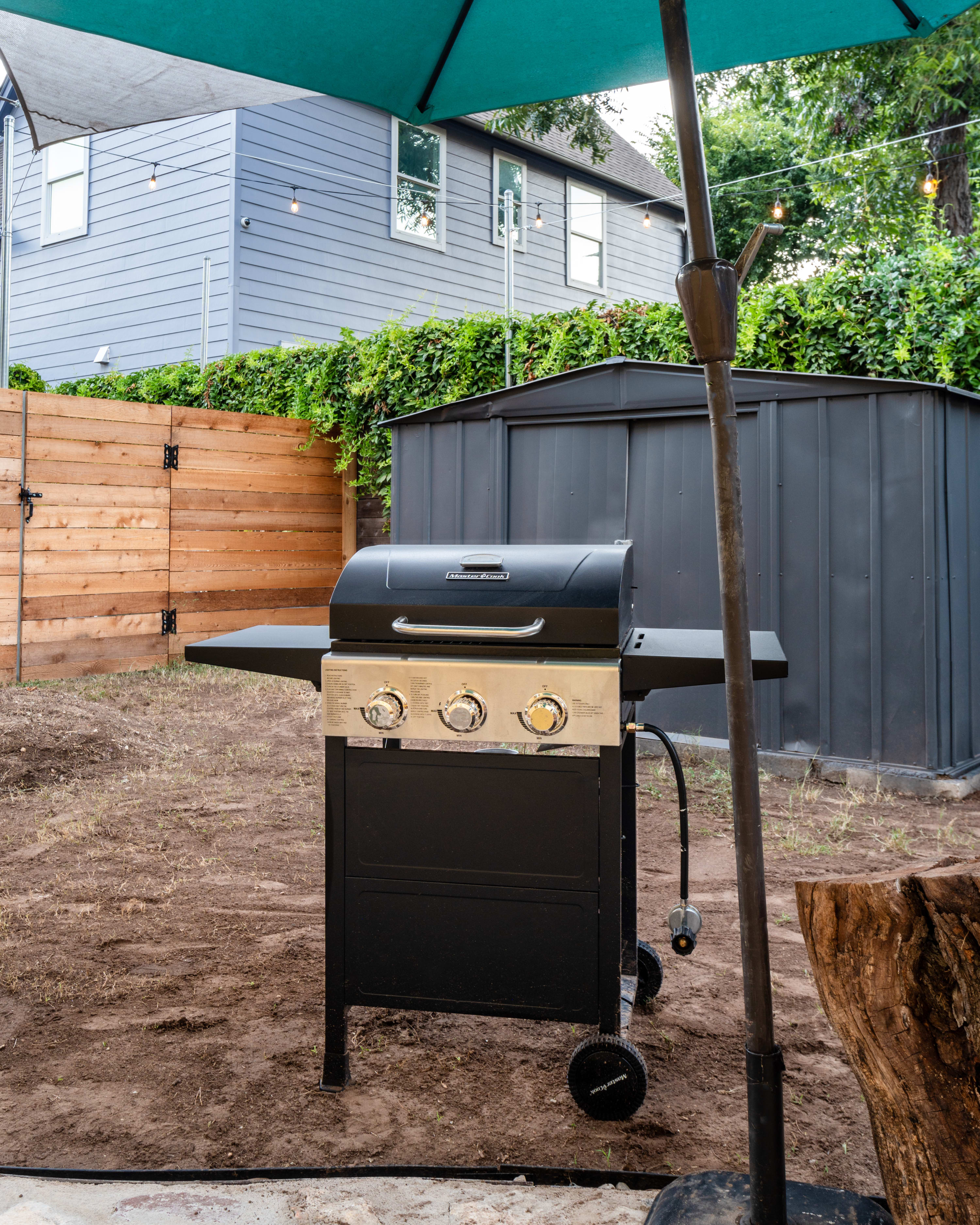 Close-up view of a black gas grill with three burners and a side table, positioned on a dirt patch in the backyard. A teal umbrella pole is visible to the right, and a shed is in the background.