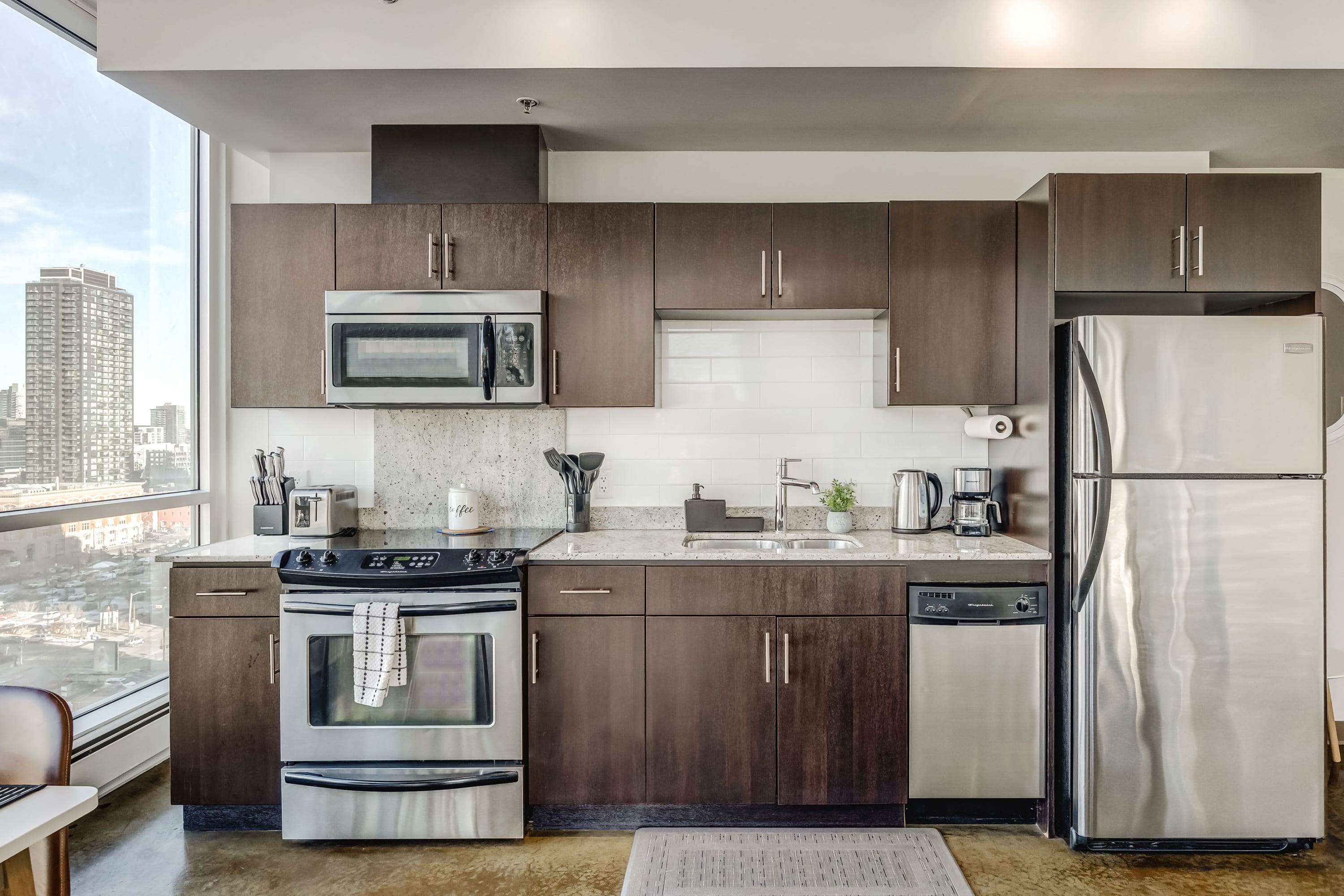 Full view of a modern kitchen featuring dark wood cabinets, light granite countertops, a white subway tile backsplash, and stainless steel appliances including a refrigerator, dishwasher, stove, and microwave. The kitchen is well-equipped with various small appliances.