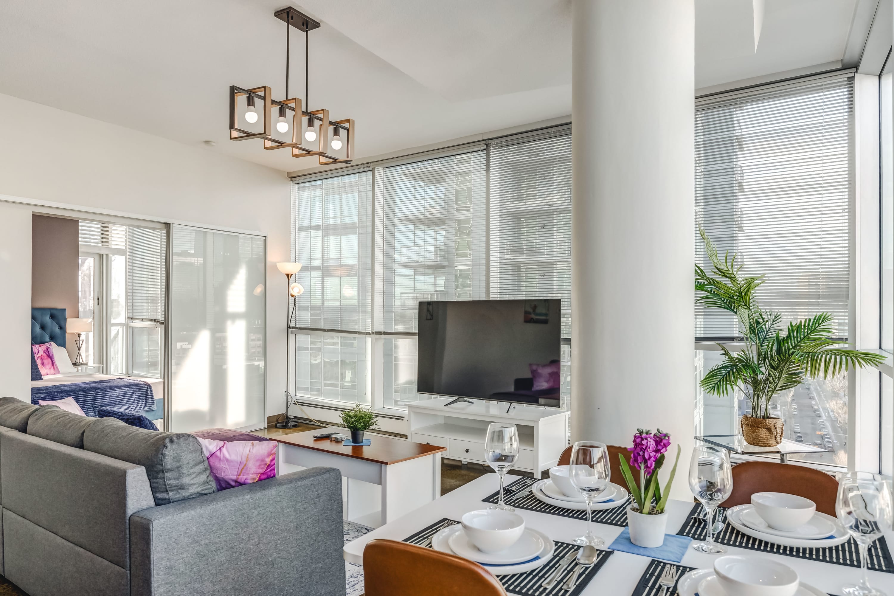 Open-concept living room with a gray sectional sofa and a dining table set for four. A modern chandelier hangs above, and a bedroom is visible through a sliding door, all complemented by large windows with city views and polished concrete floors.